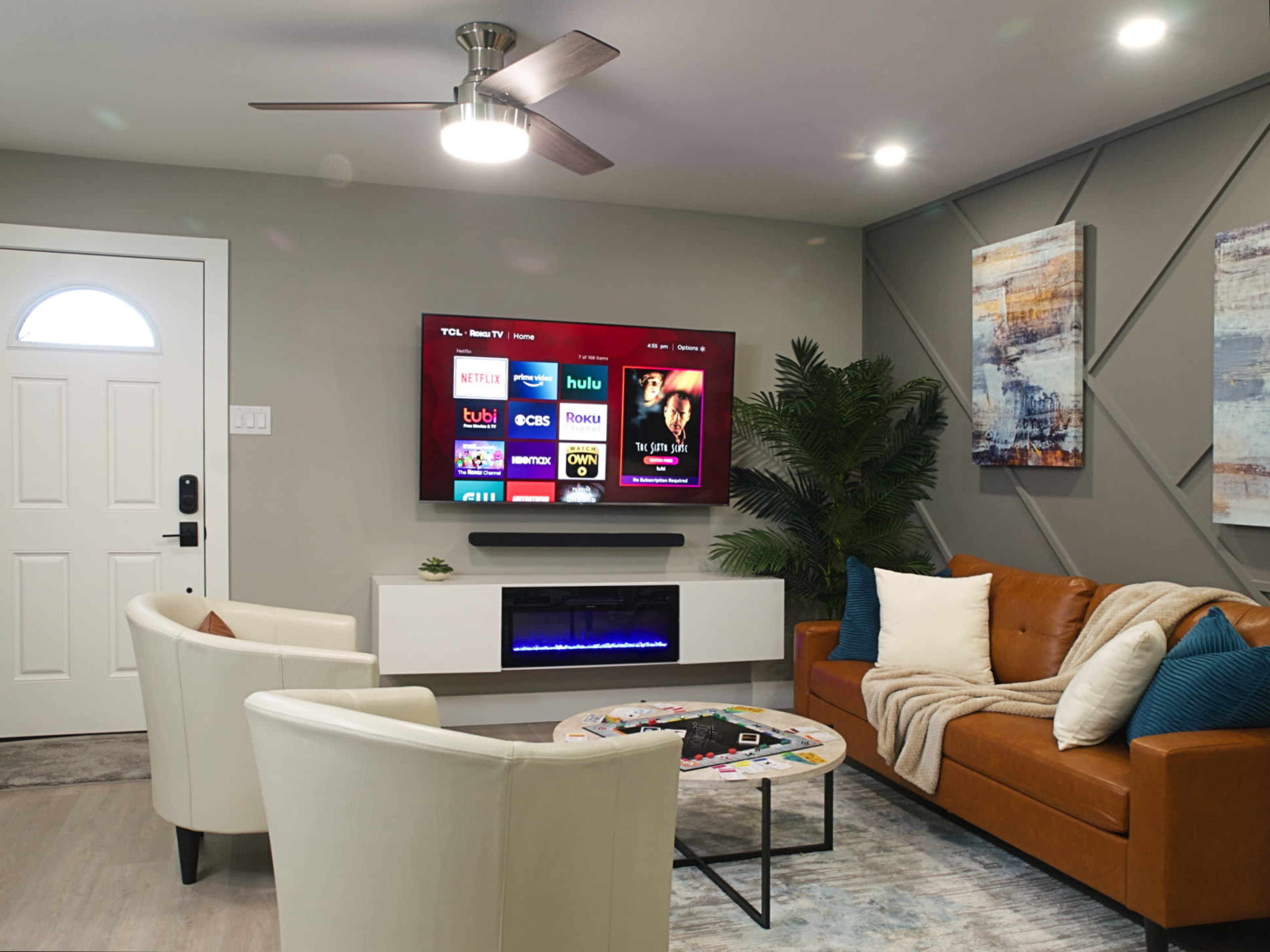 A modern living room features a leather sofa, two white armchairs, a wall-mounted TV displaying streaming options, and a decorative coffee table in front.