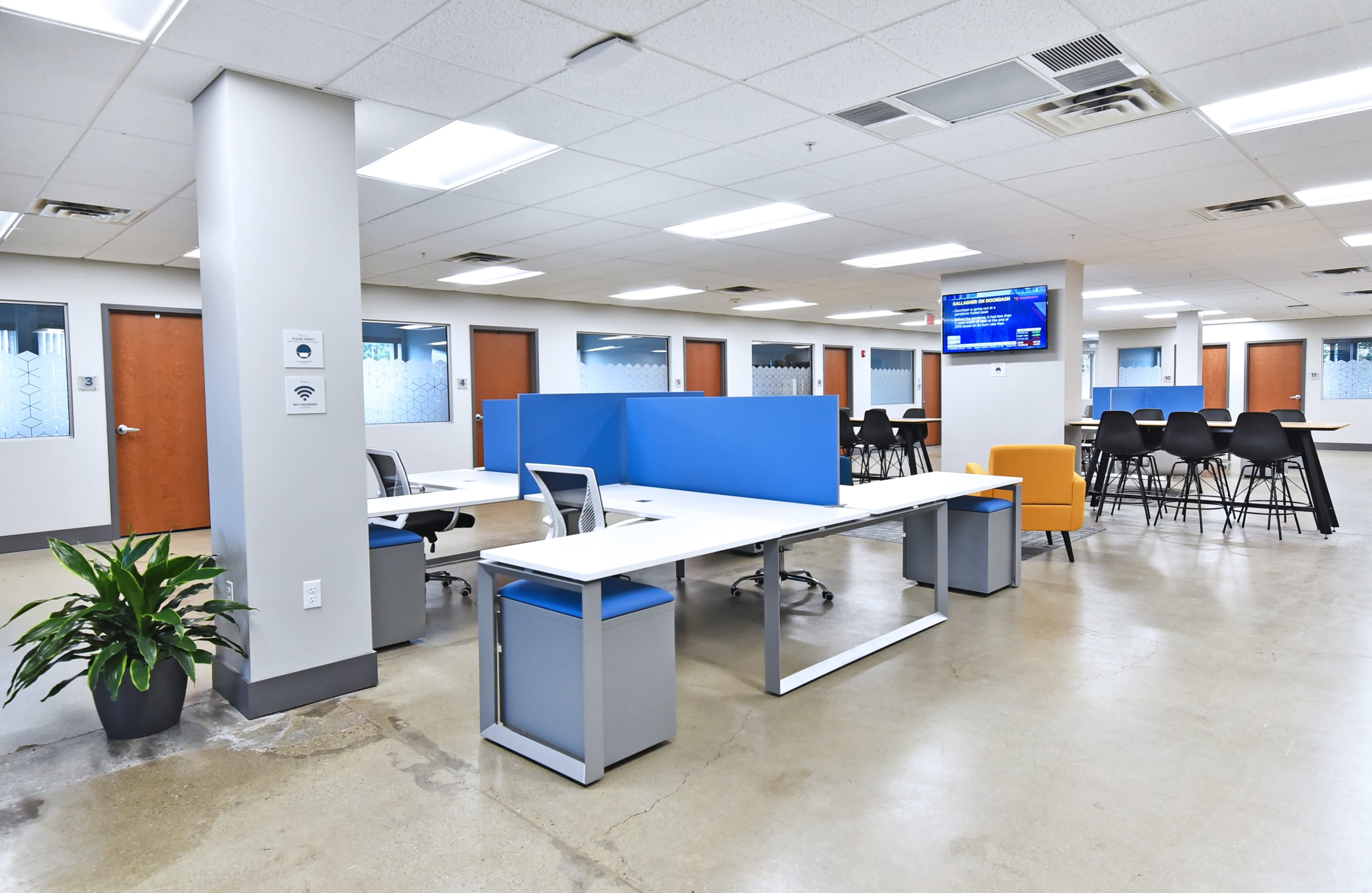 The image shows a modern office space with several workstations, a seating area, and a digital screen displaying information.