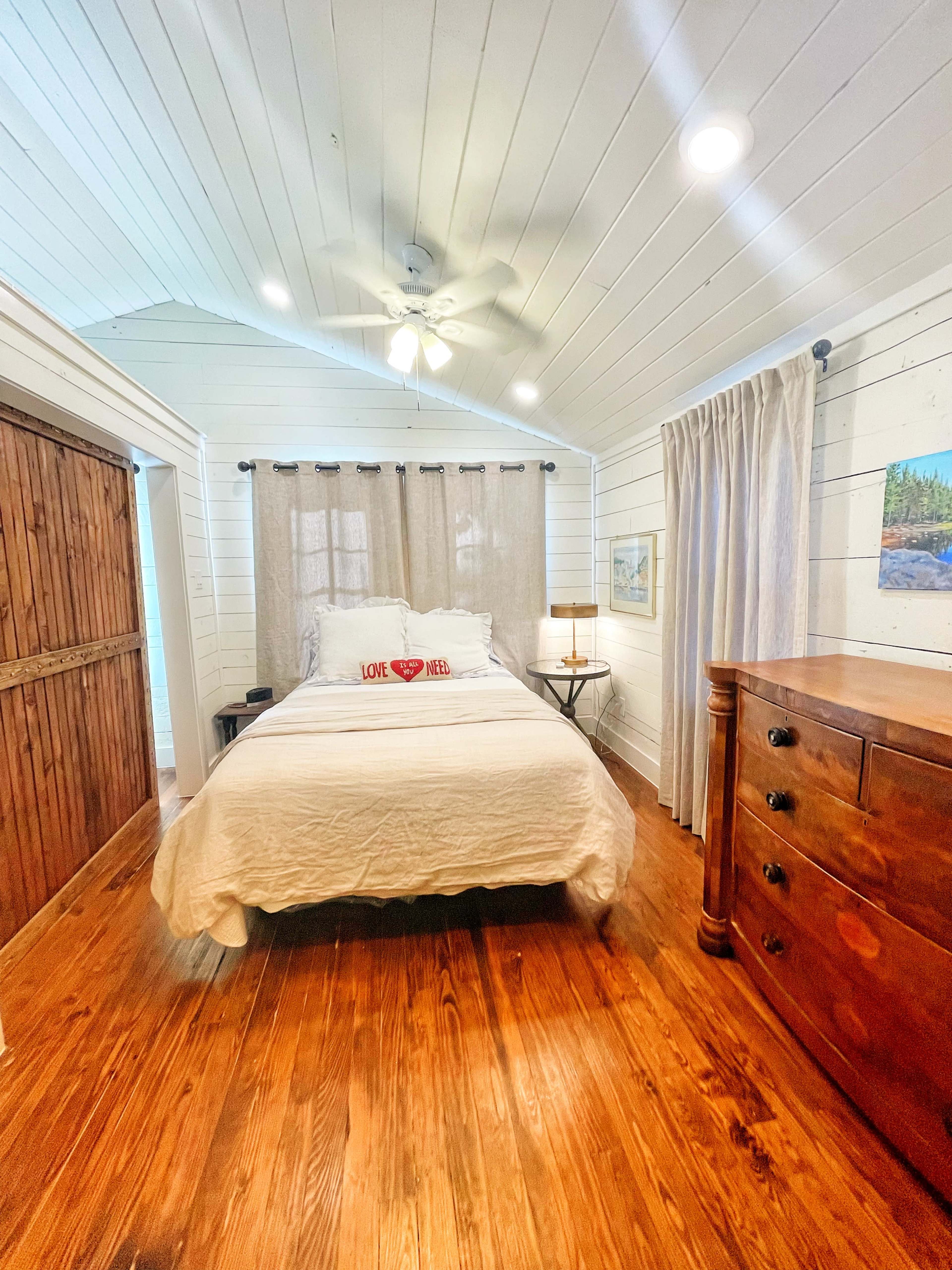 A cozy bedroom features a bed with white bedding, wooden flooring, a ceiling fan, and light-colored curtains.