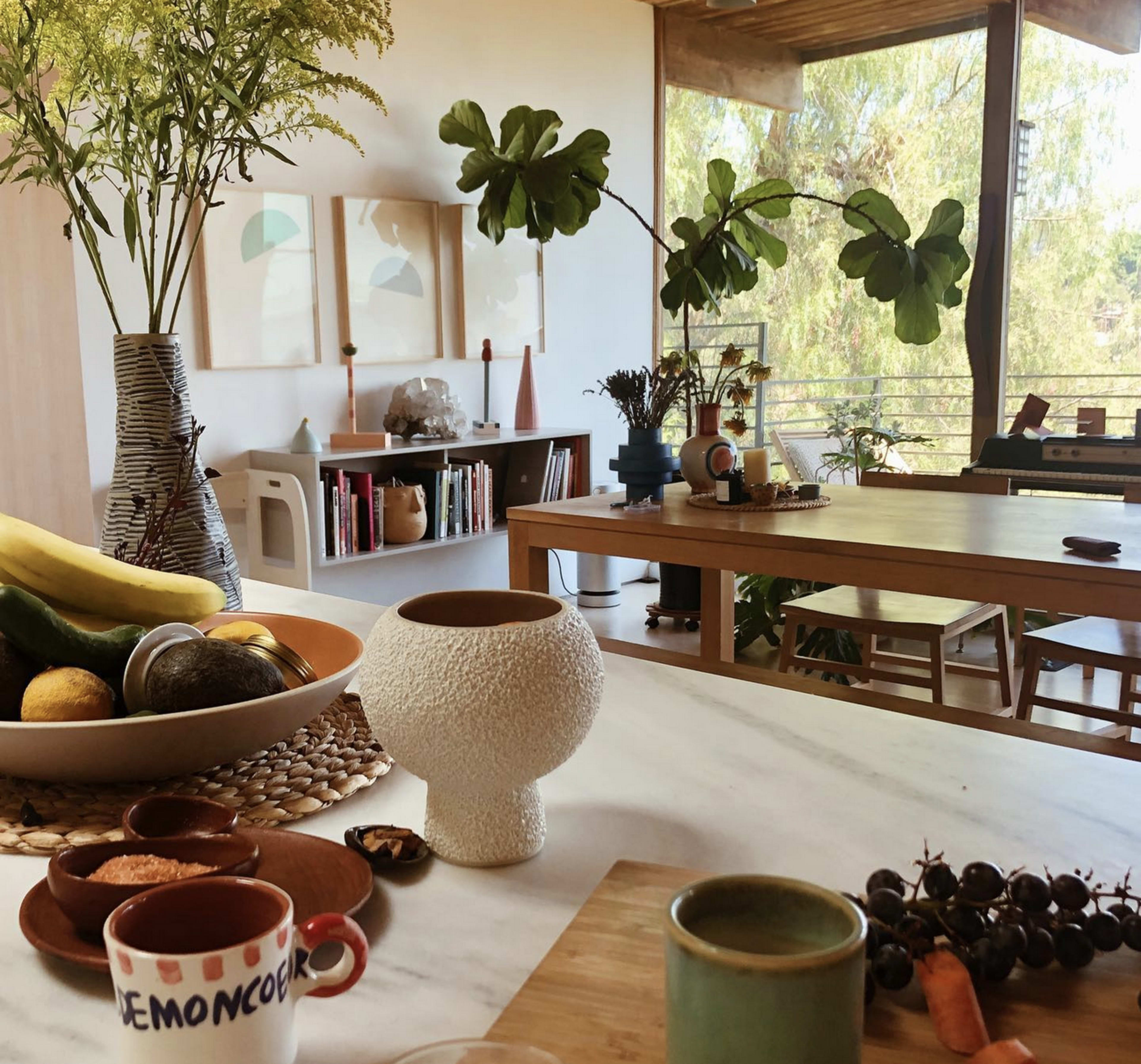 A modern interior features a dining table with fruit, a textured vase, and large windows that open to a view of greenery.
