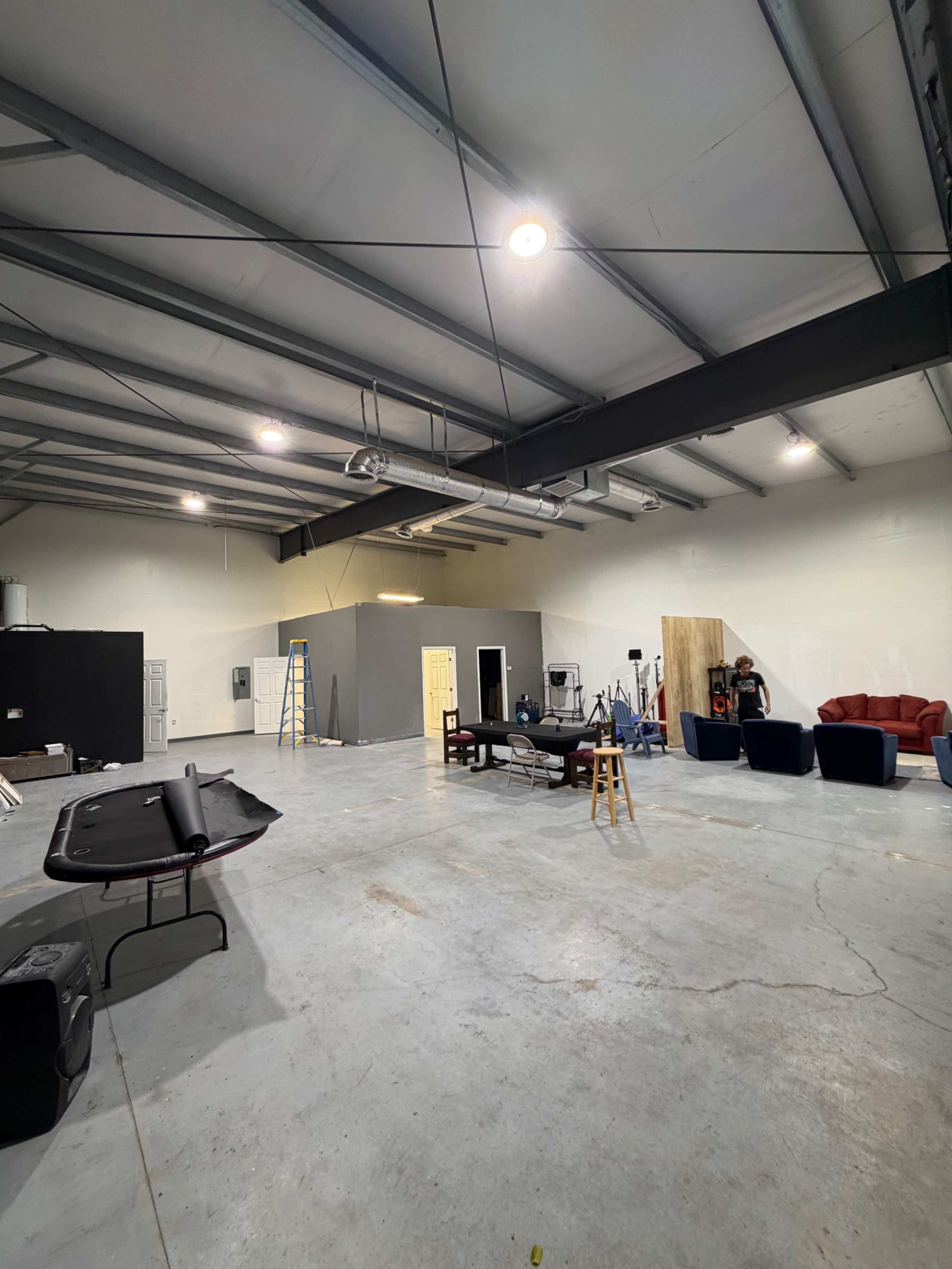 The image shows an empty industrial-style studio space with exposed beams, concrete flooring, and various furniture and equipment scattered around.