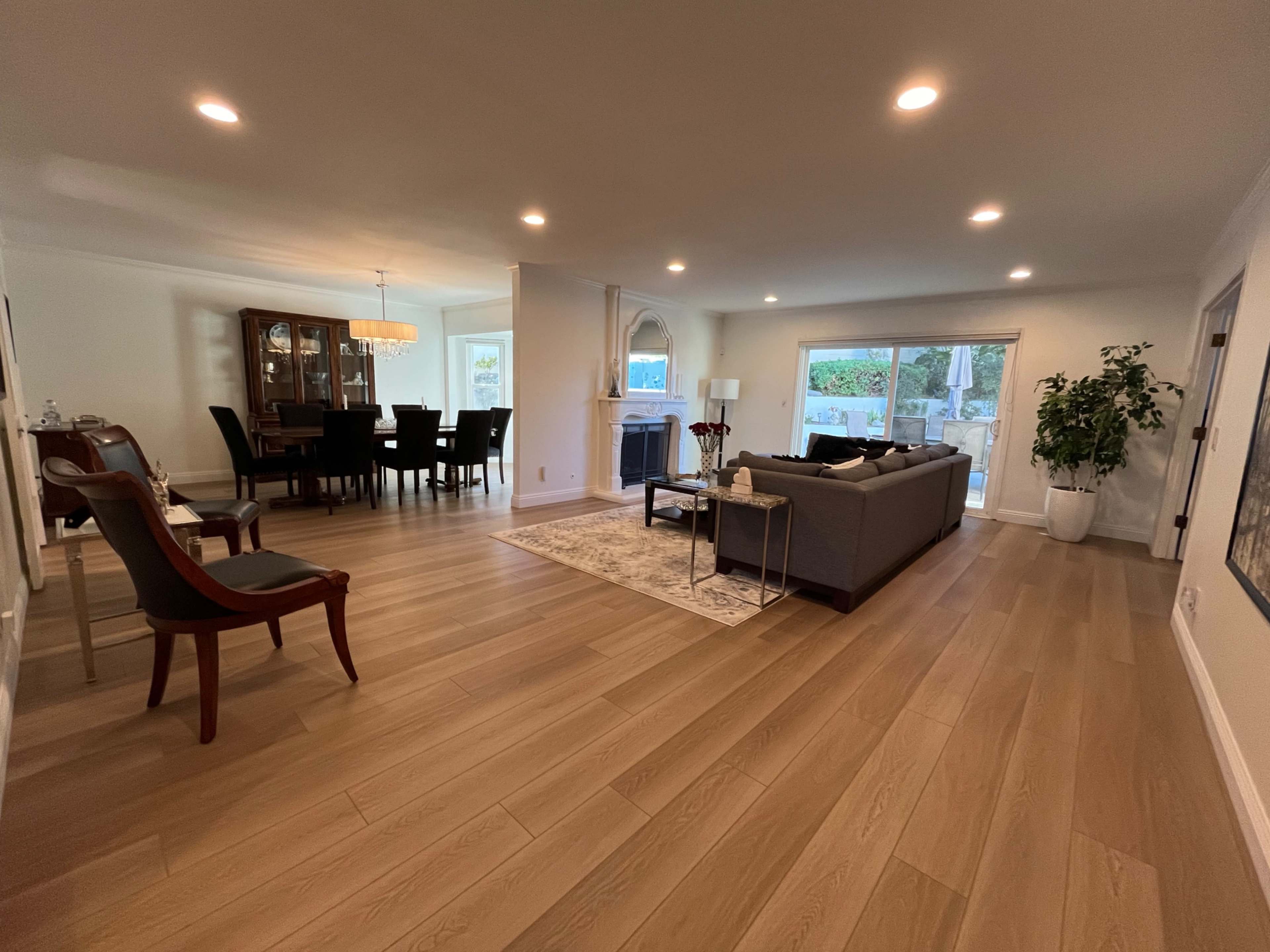 The image shows a spacious living room with hardwood floors, featuring a dining area in the background, a large sectional sofa, and a glass door leading to an outdoor space.