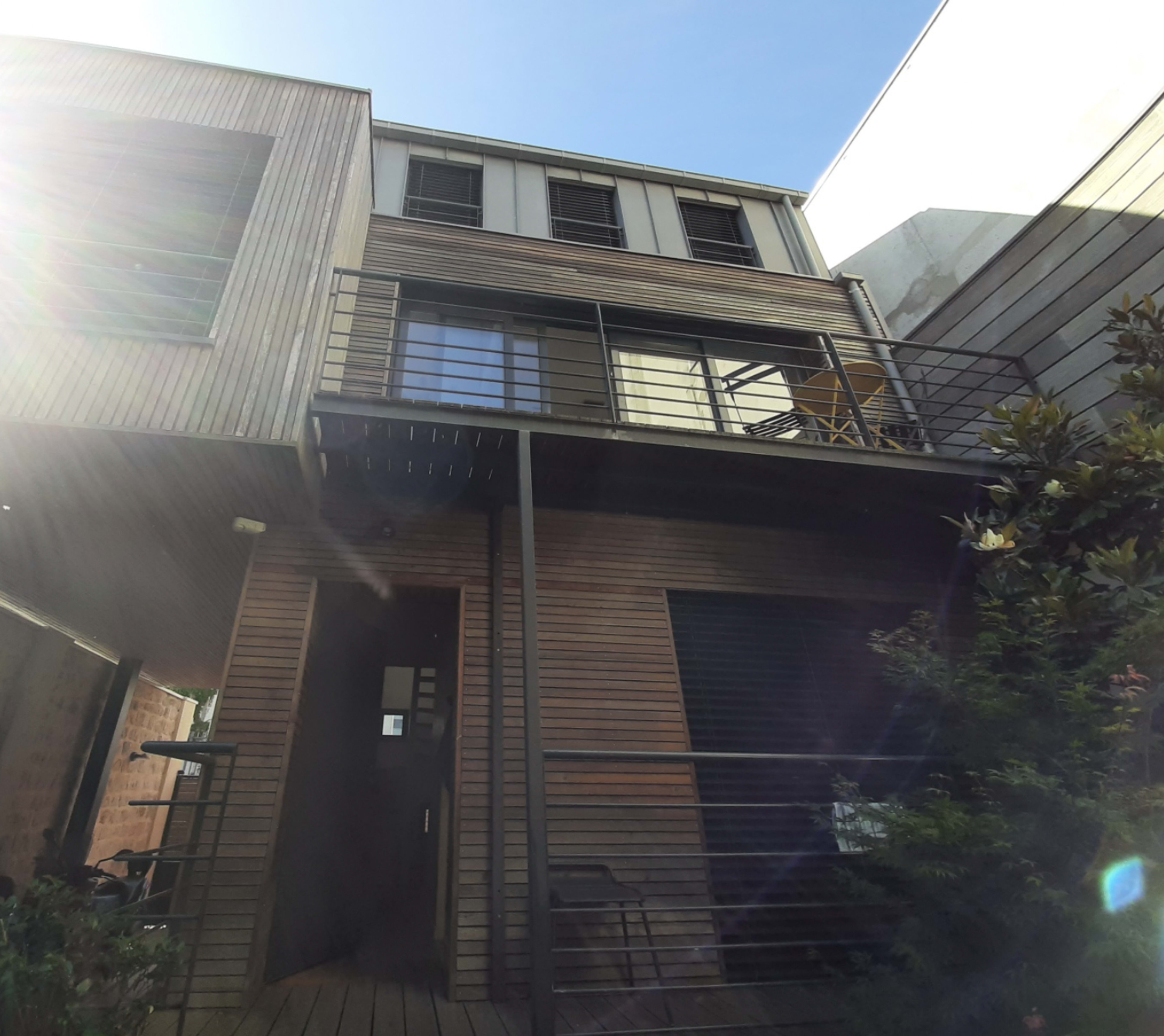 A two-story wooden structure with a balcony and a door at the ground level, surrounded by greenery under a clear blue sky.