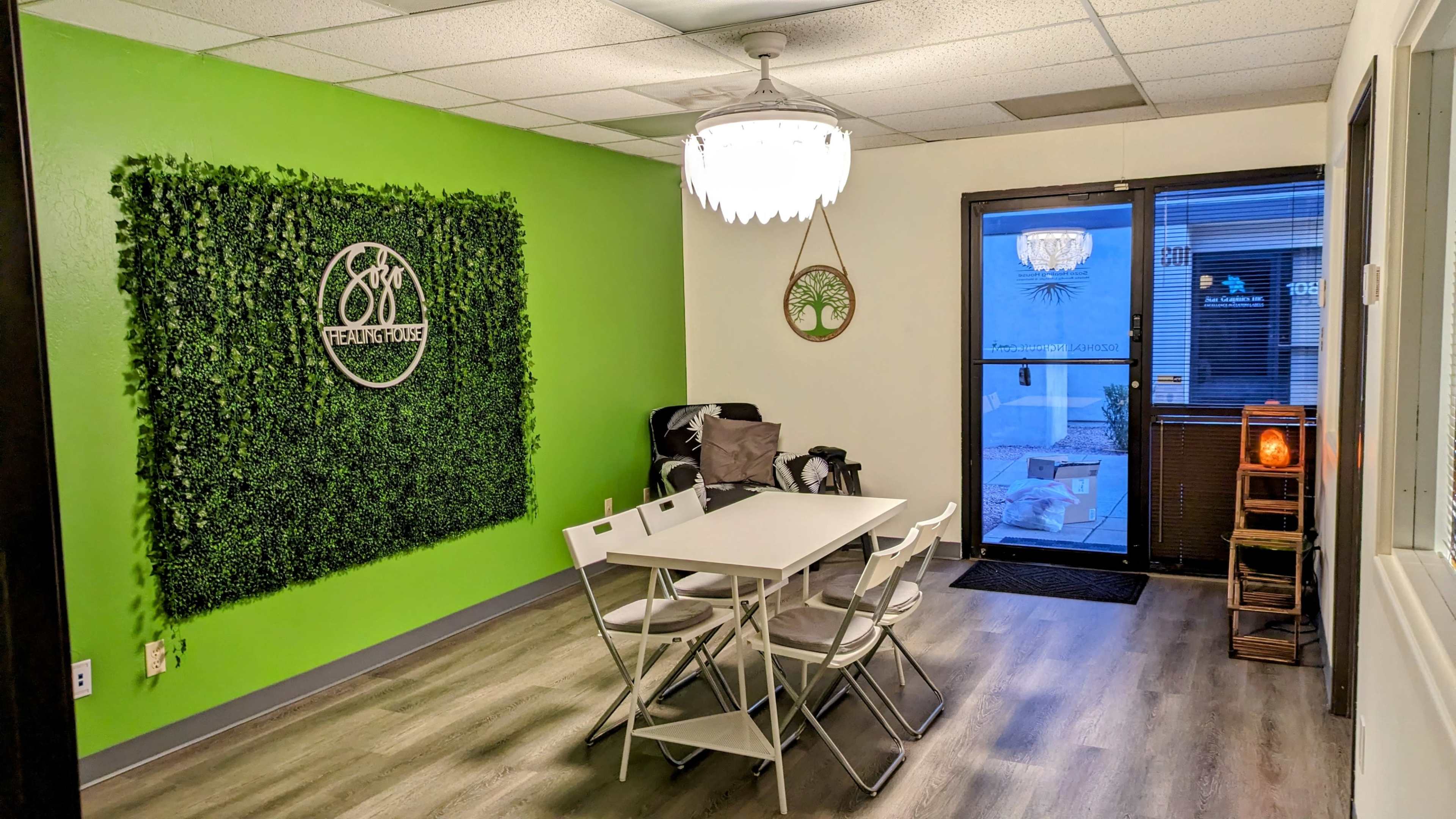 A modern room with a green accent wall featuring a plant wall decor, a white table surrounded by chairs, and large windows allowing natural light.