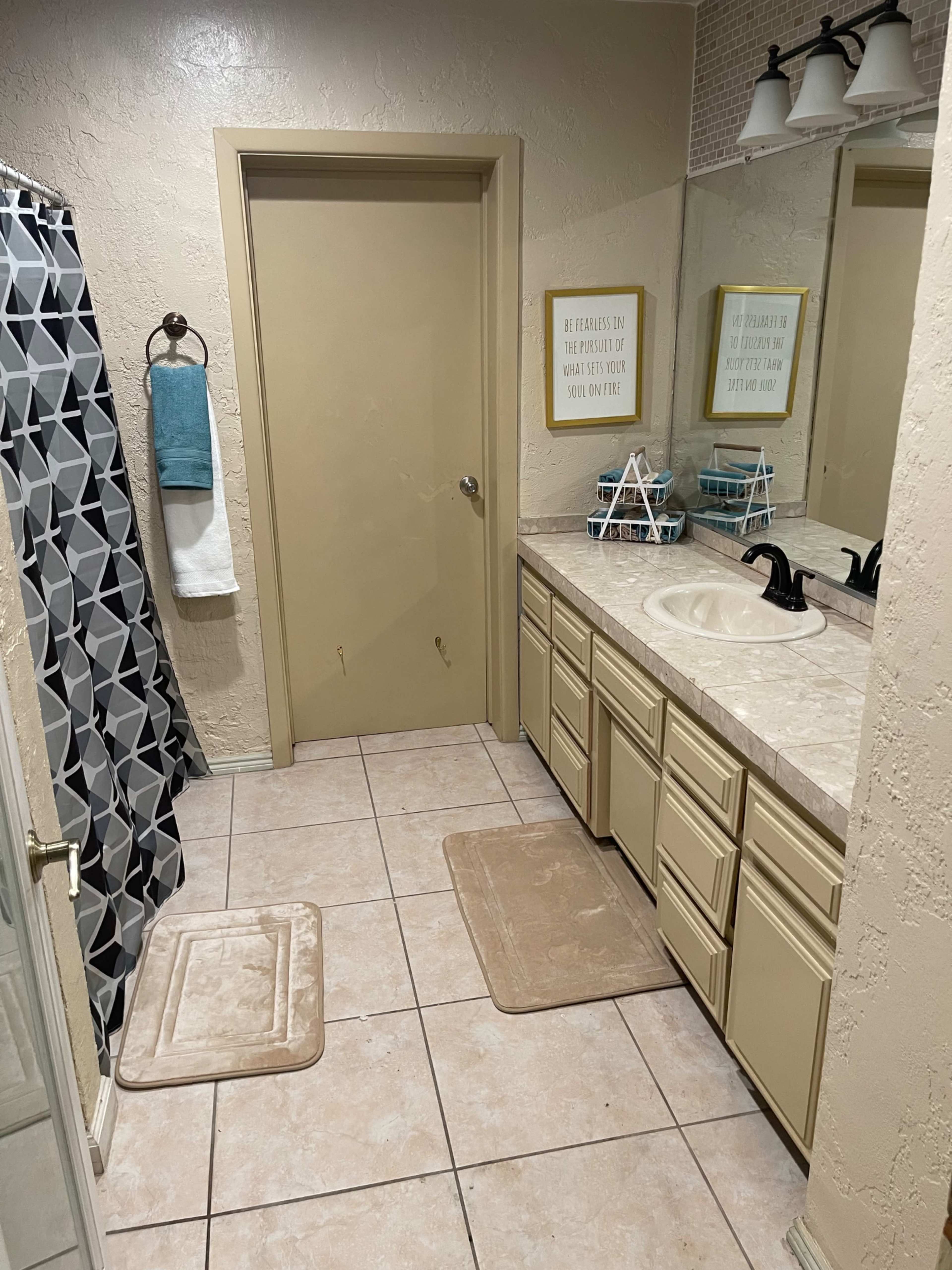 A bathroom with a sink area, a shower curtain, and a door, featuring beige cabinetry and tiled flooring.