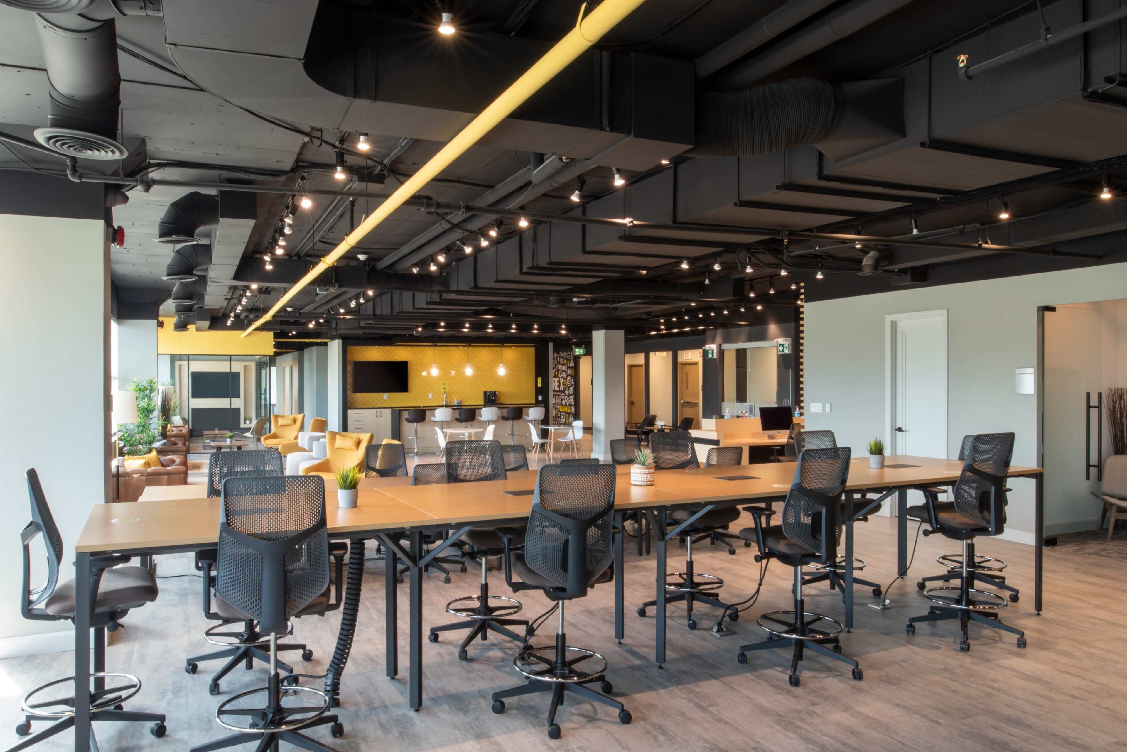 A modern office space with a large communal table surrounded by tall chairs, features bright overhead lighting, and various seating arrangements in the background.
