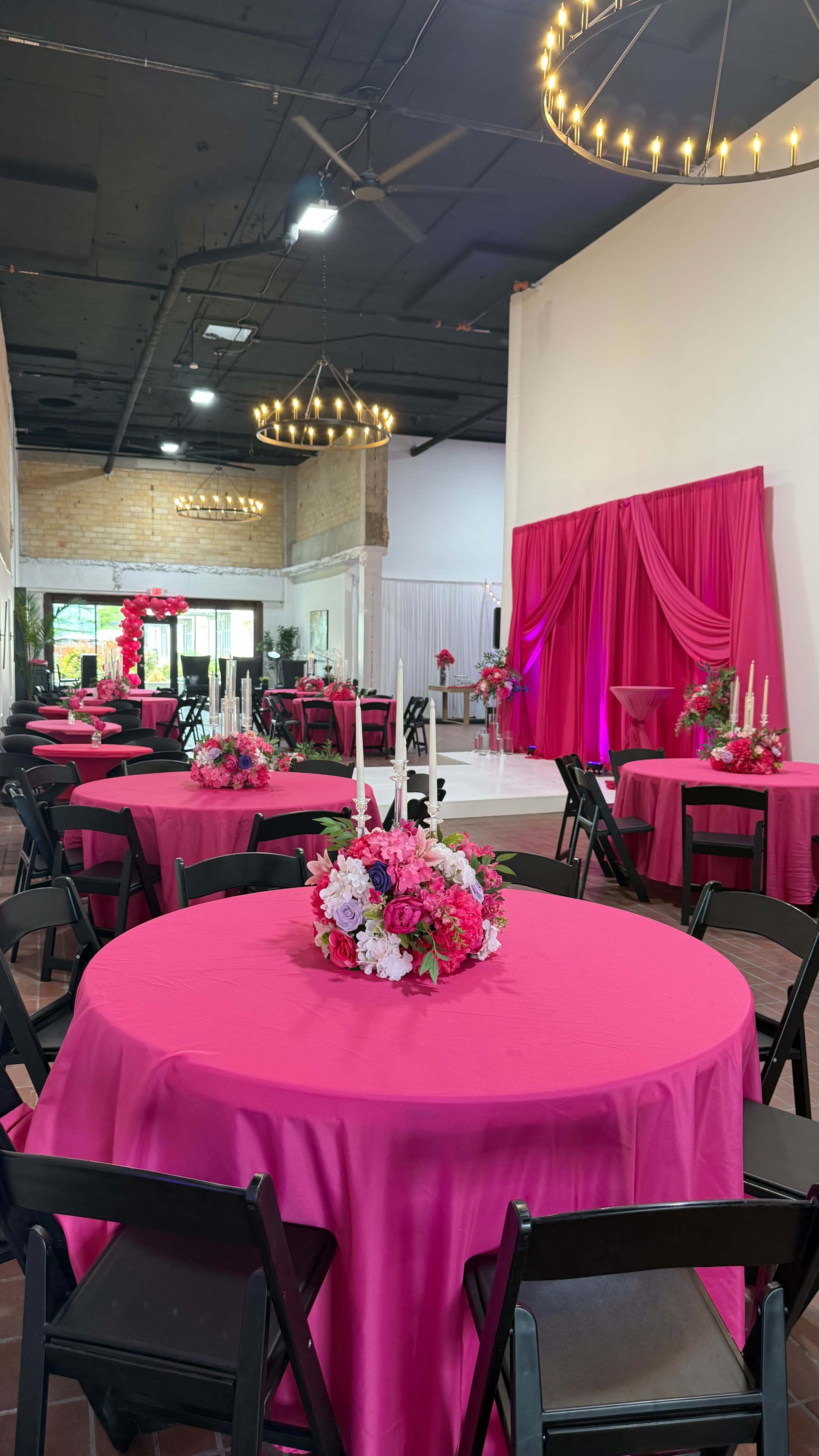 The image shows a vibrant event space decorated with pink tablecloths, floral centerpieces, and a backdrop draped in pink fabric.