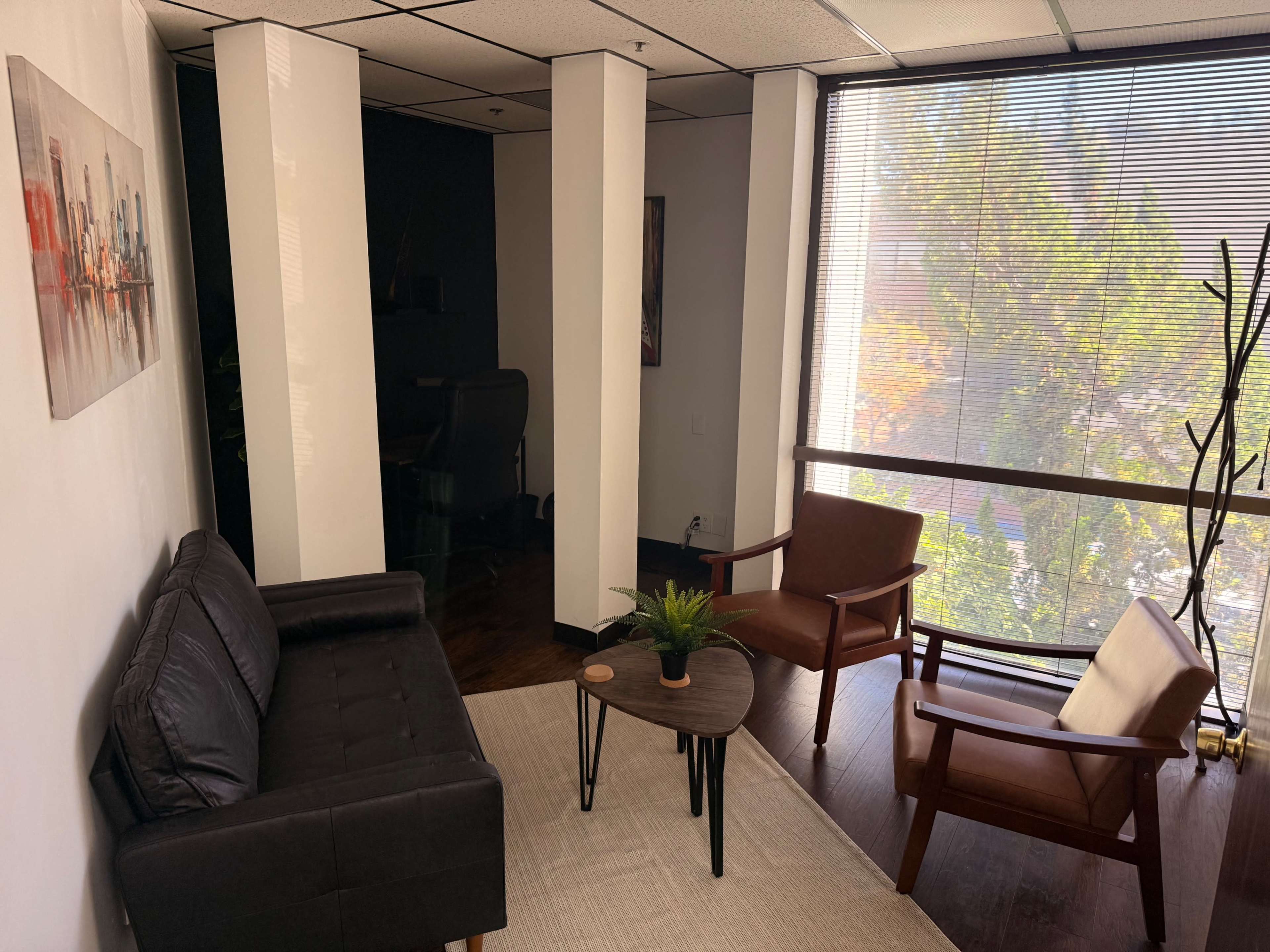 The image depicts a small, modern office space with a black couch, two wooden chairs, a round table, and a large window allowing natural light to enter.