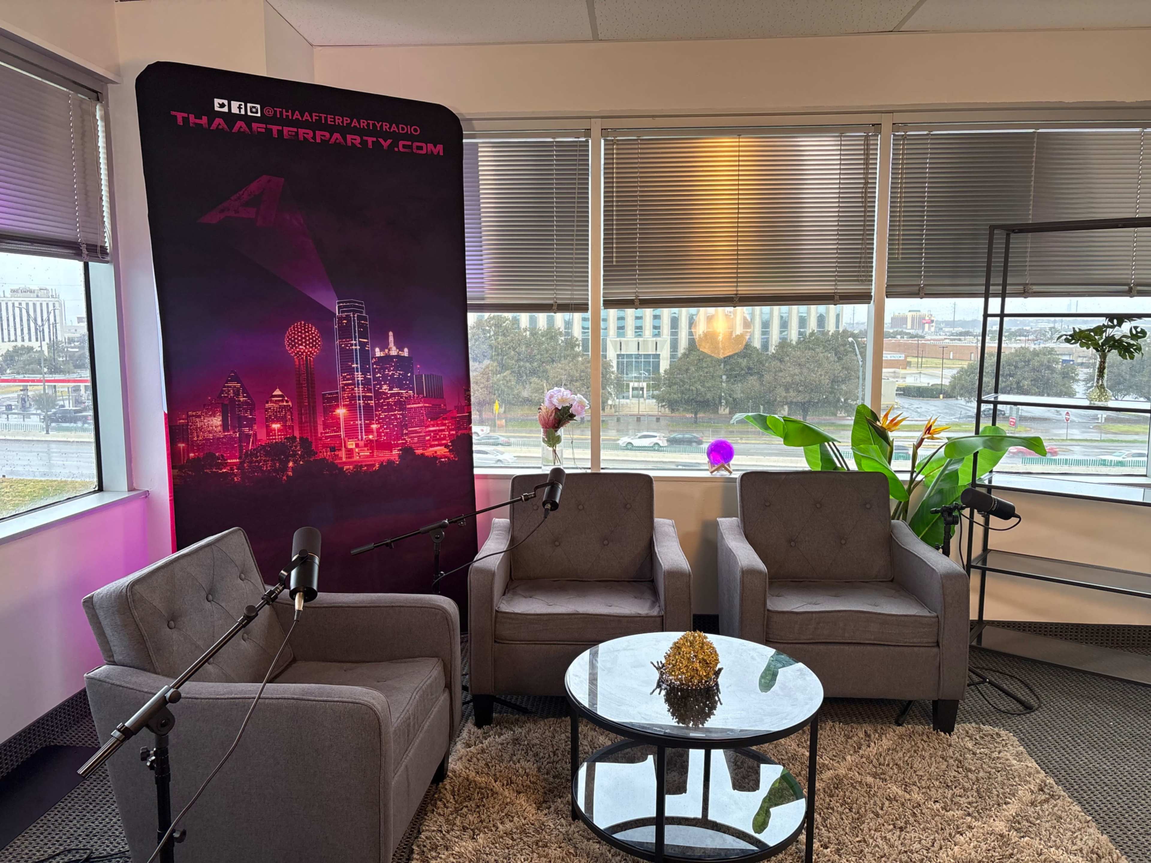 The image shows a cozy interview setup with two gray armchairs facing a small glass table, accompanied by microphones and a backdrop featuring city skyline graphics and a website.