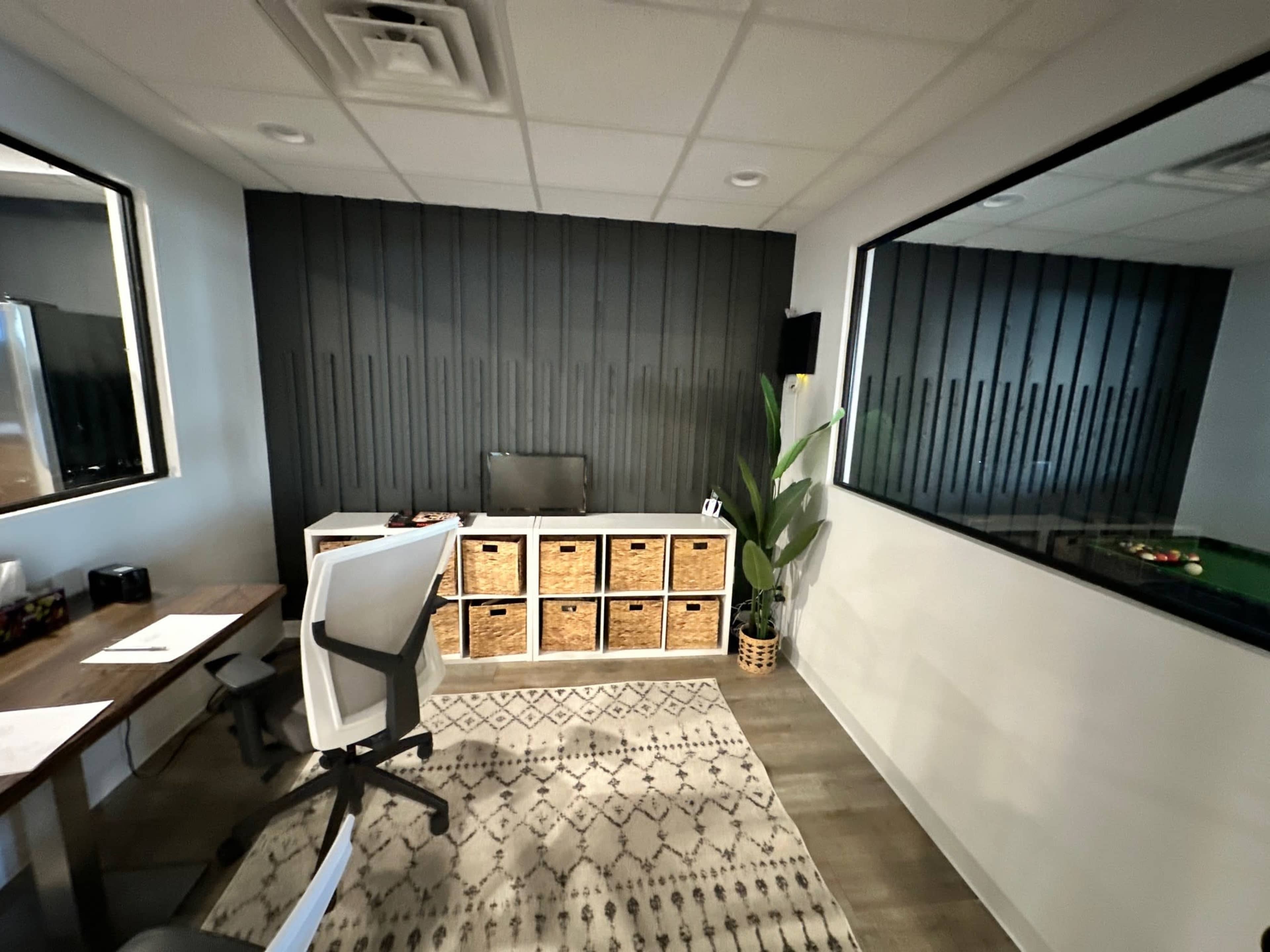 A small office space with a desk, a chair, storage boxes on a shelf, and a large window overlooking another room.