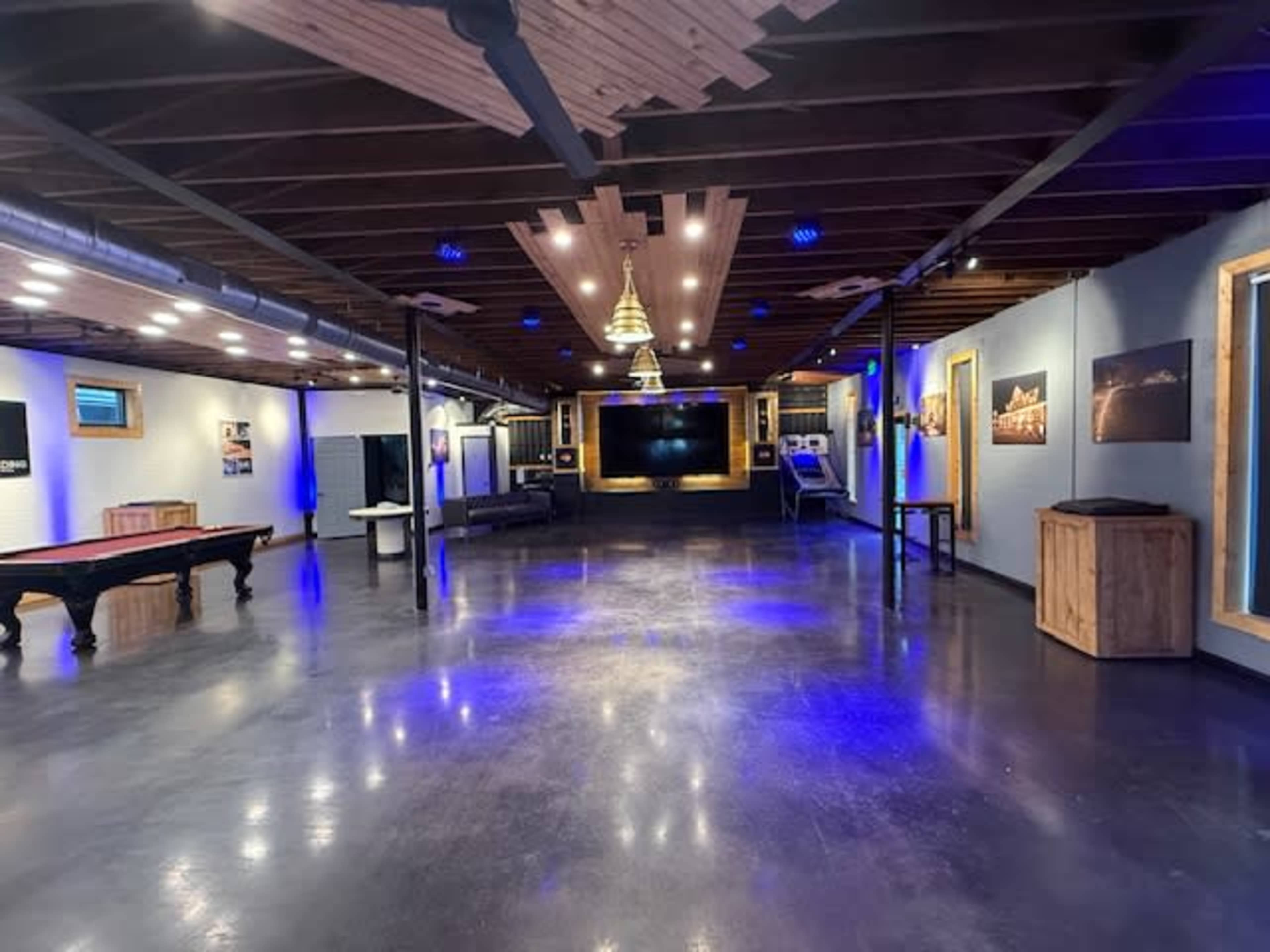The image shows a spacious interior with polished concrete floors, wooden beams, and blue ambient lighting, featuring a large screen, seating areas, and a billiard table.