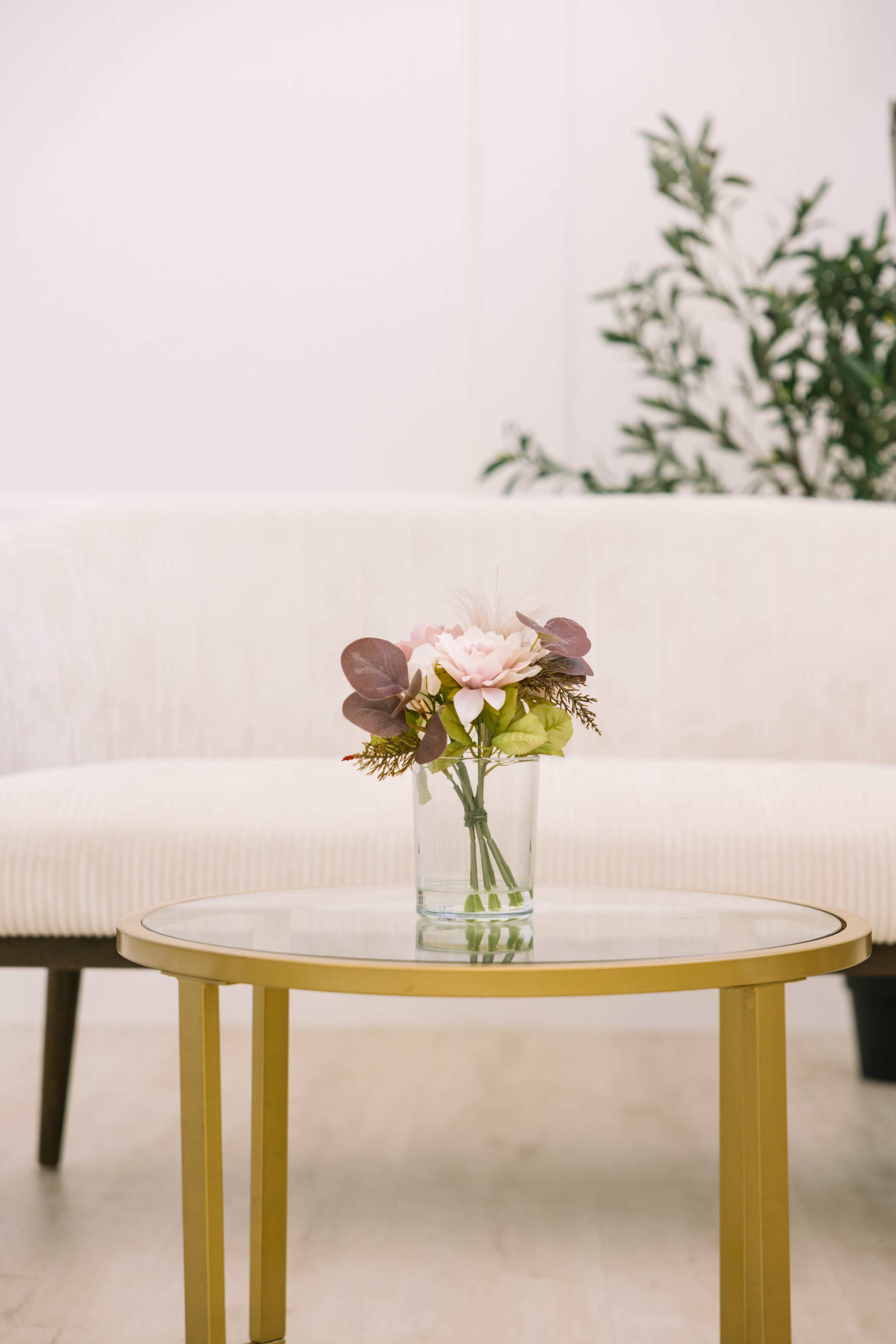 A glass vase with flowers sits on a gold coffee table in front of a light-colored couch.