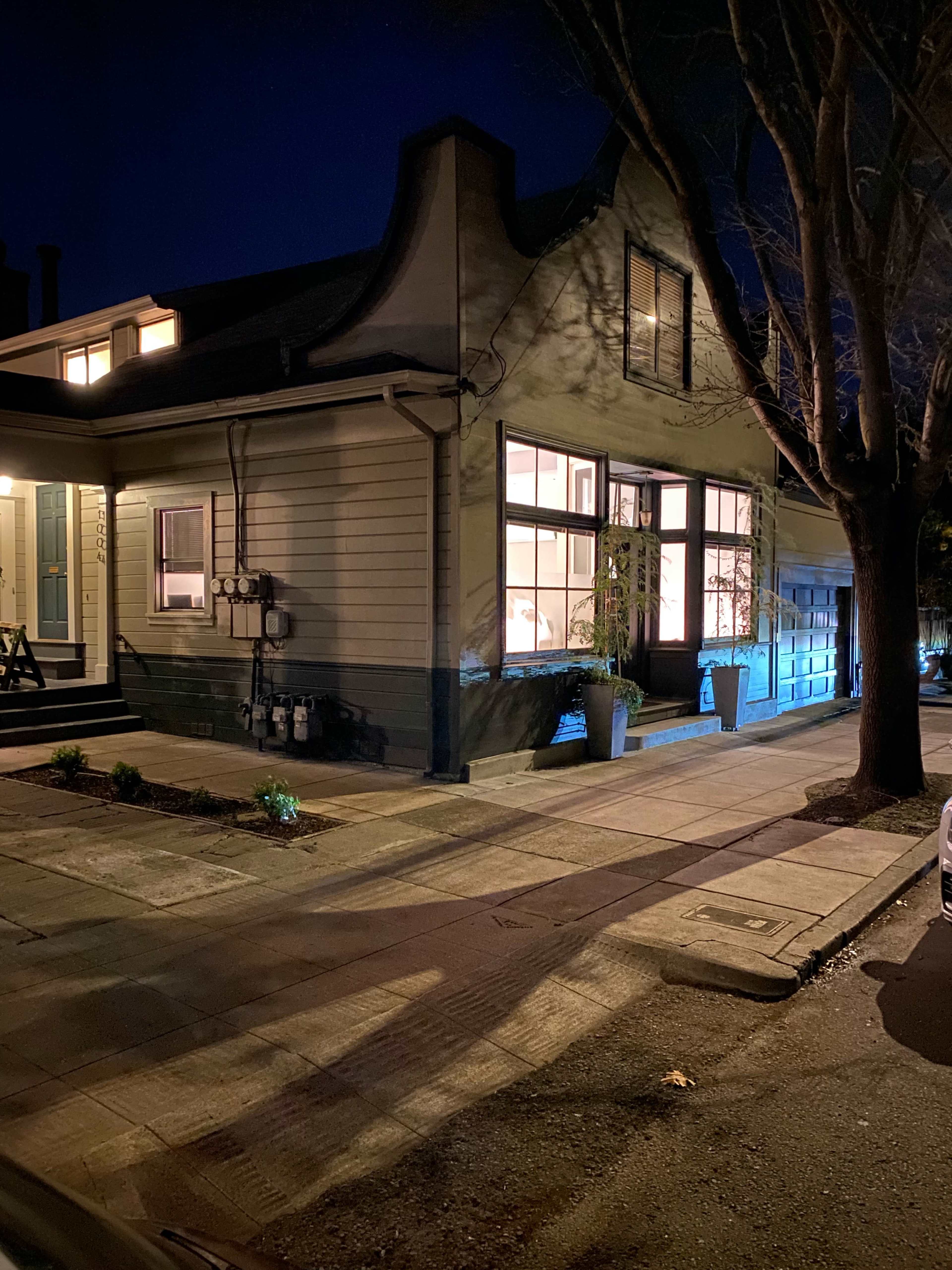 A house with a distinctive roofline is illuminated at night, showcasing windows that reflect warm light, while a tree casts shadows on the sidewalk.