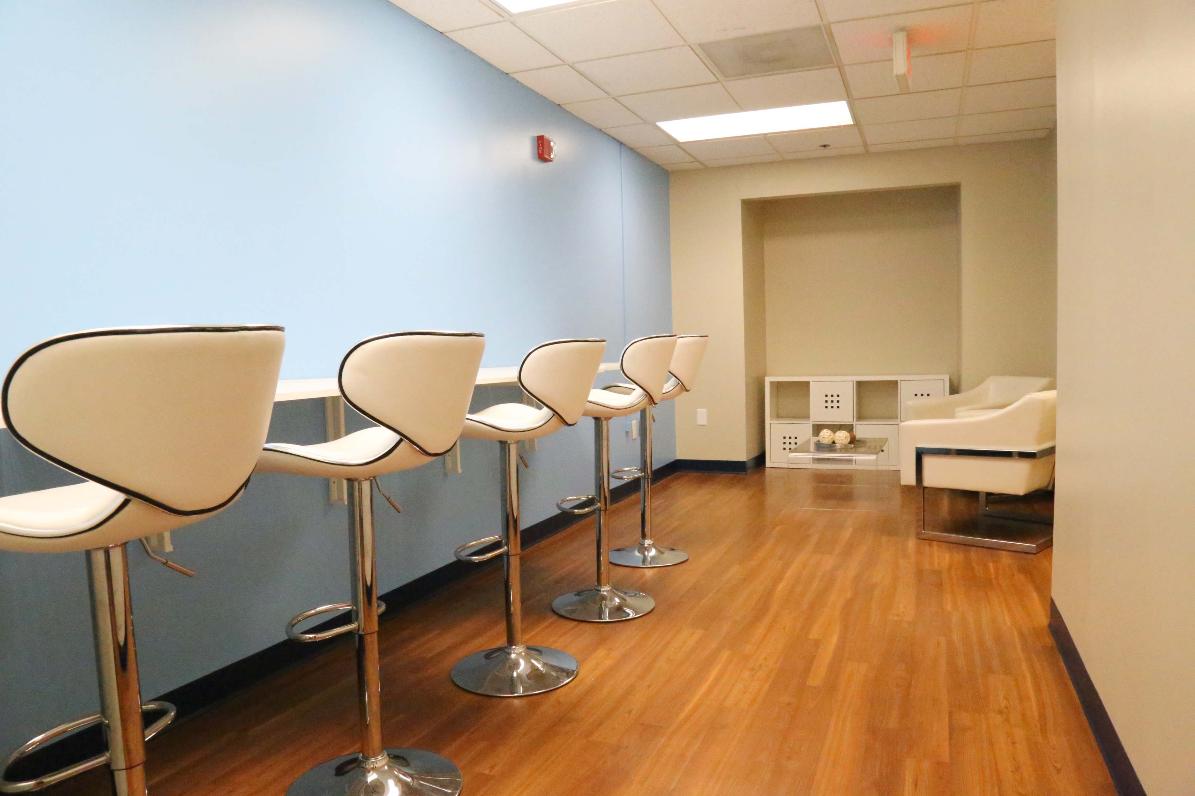 A modern lounge area with several white bar stools lined against a light blue wall, and a small seating area with a white chair in the background.