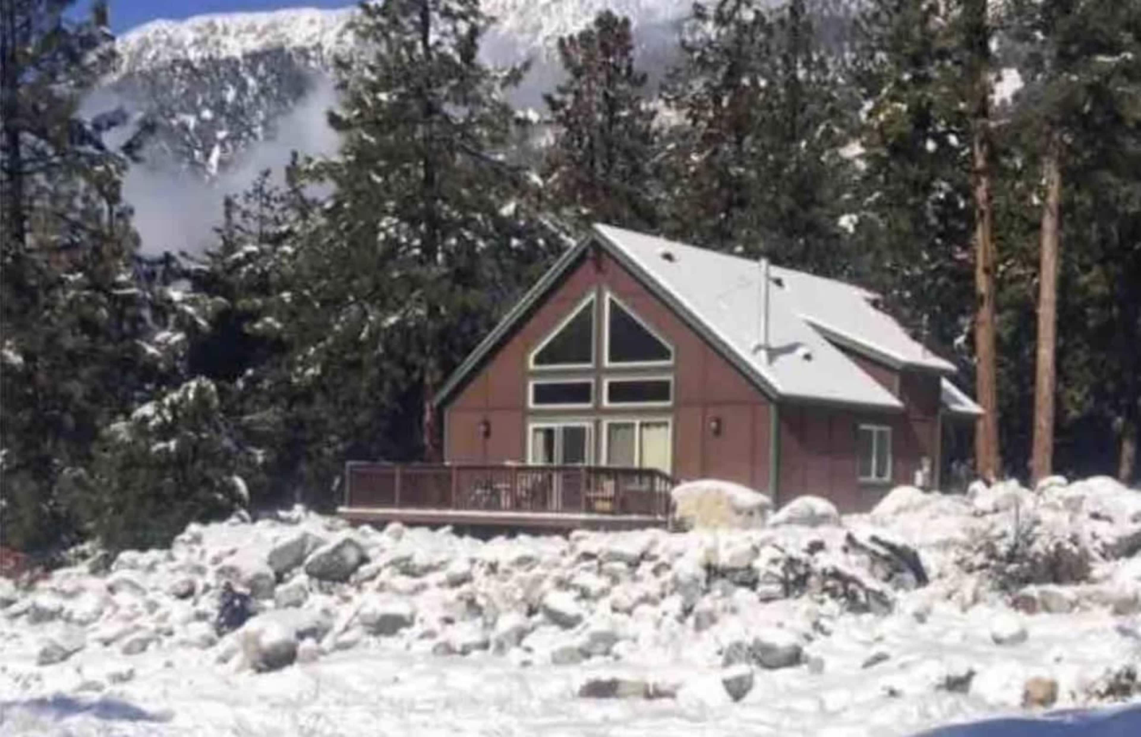 A cabin sits surrounded by snow and large rocks, with a forested area and mountains in the background.