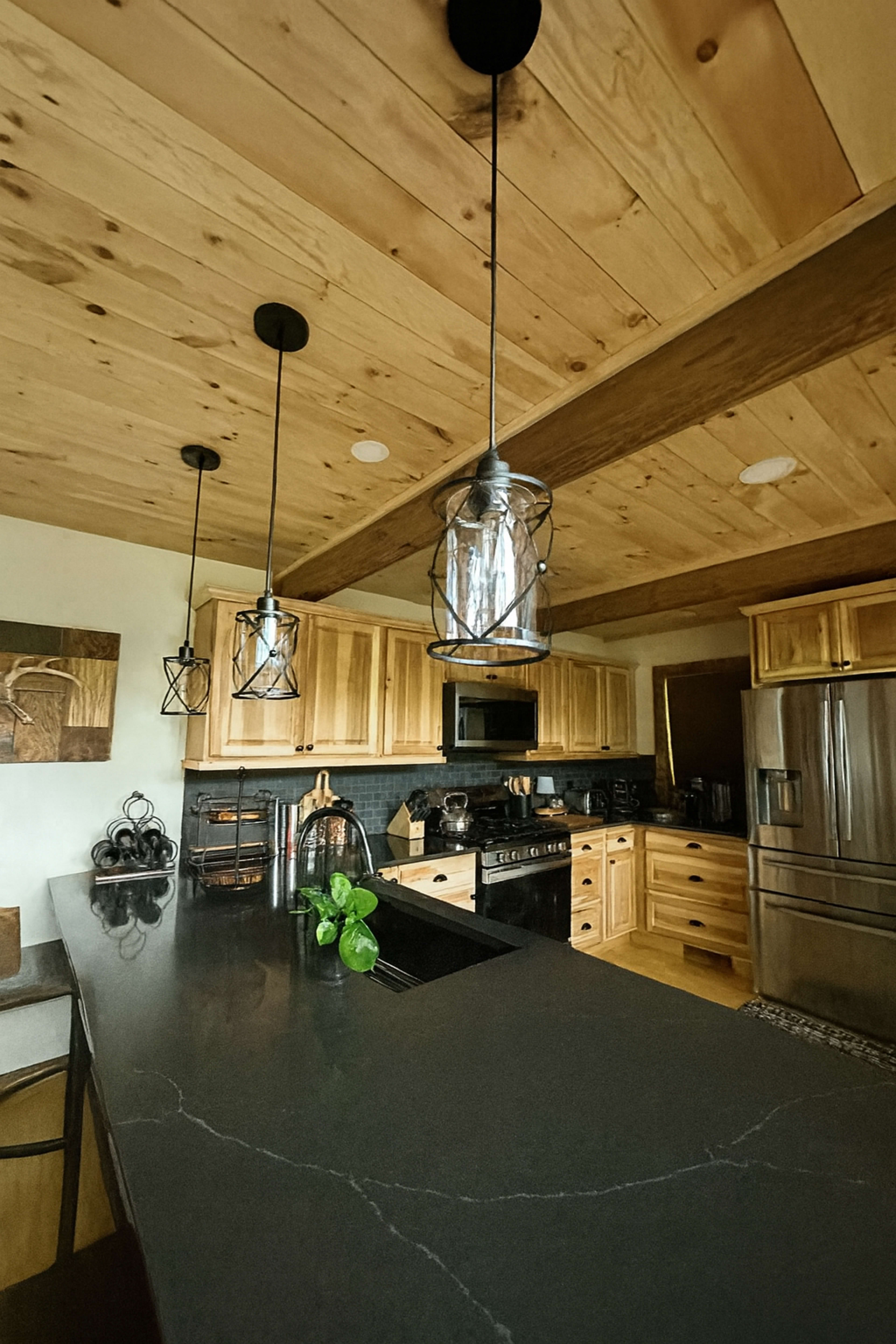 The image shows a modern kitchen with wooden cabinetry, black countertops, and three pendant lights hanging from a wooden ceiling.