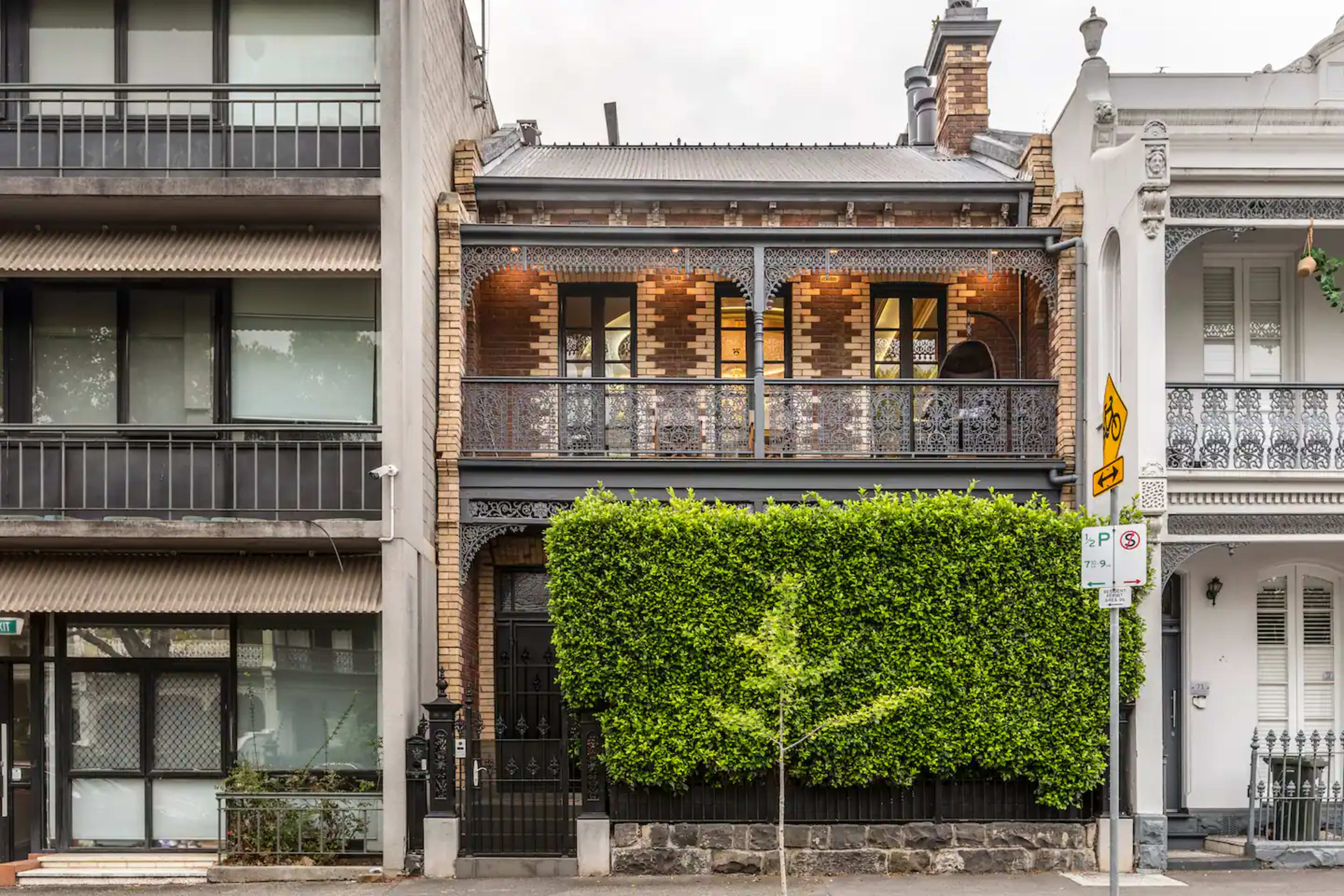 A two-story brick house with a balcony is partially obscured by a tall hedge, situated between two modern structures.