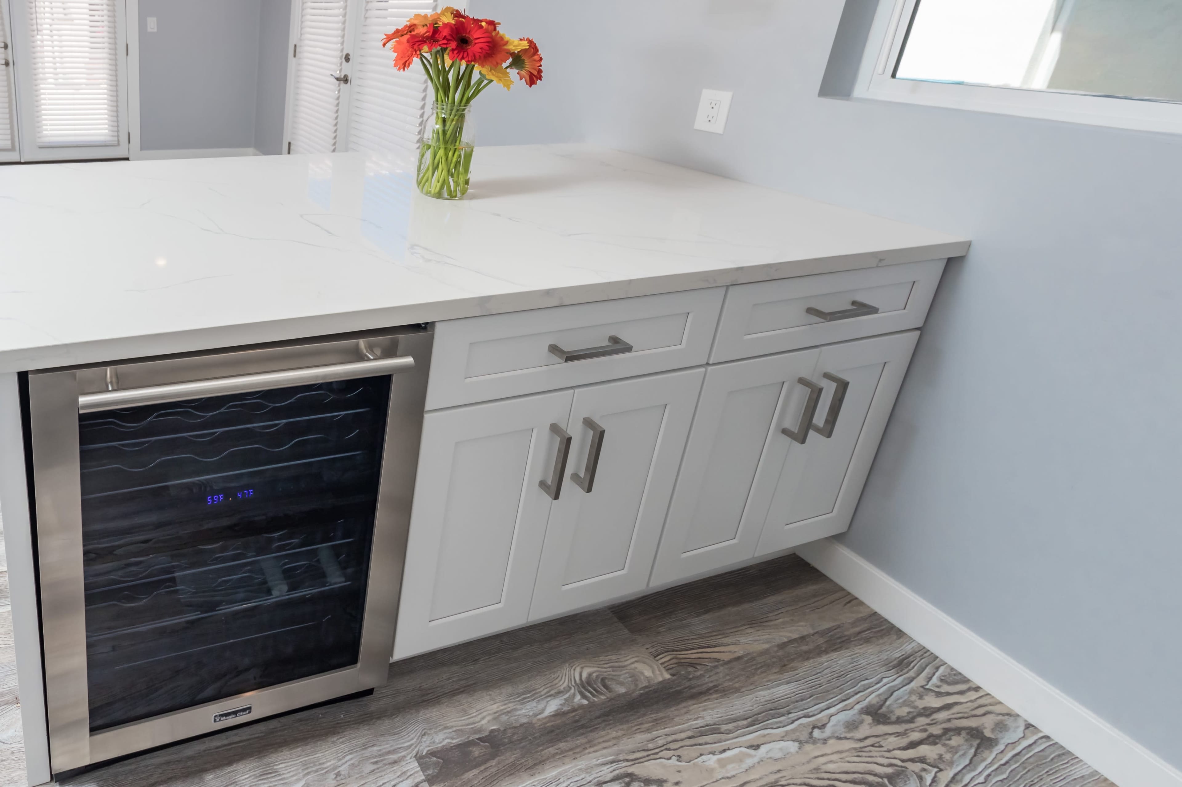 A modern kitchen features a white marble countertop with a stainless steel wine cooler and a vase of flowers on top.