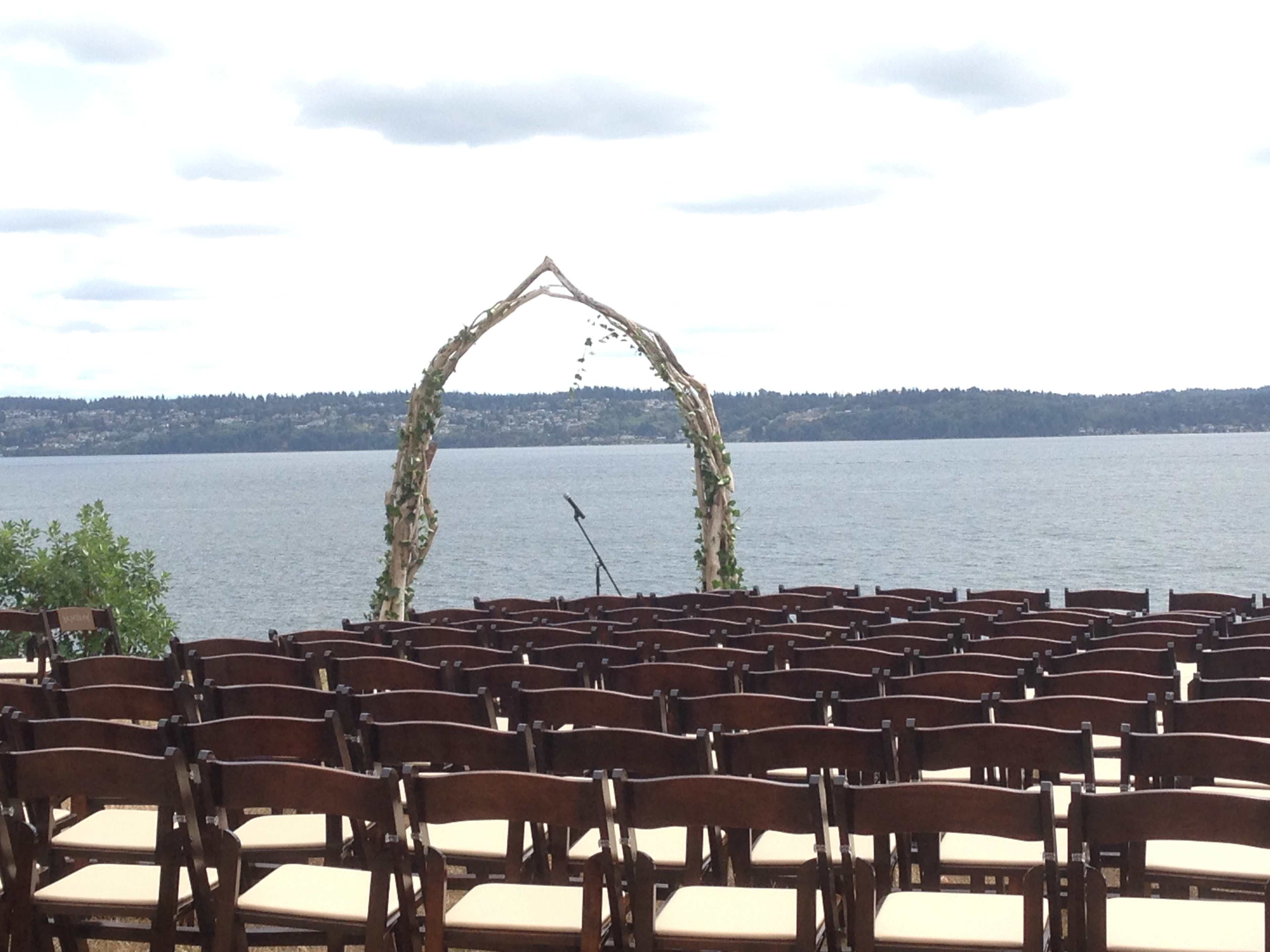 A wedding altar made of wooden branches is set up by a lake, with rows of empty chairs facing it.