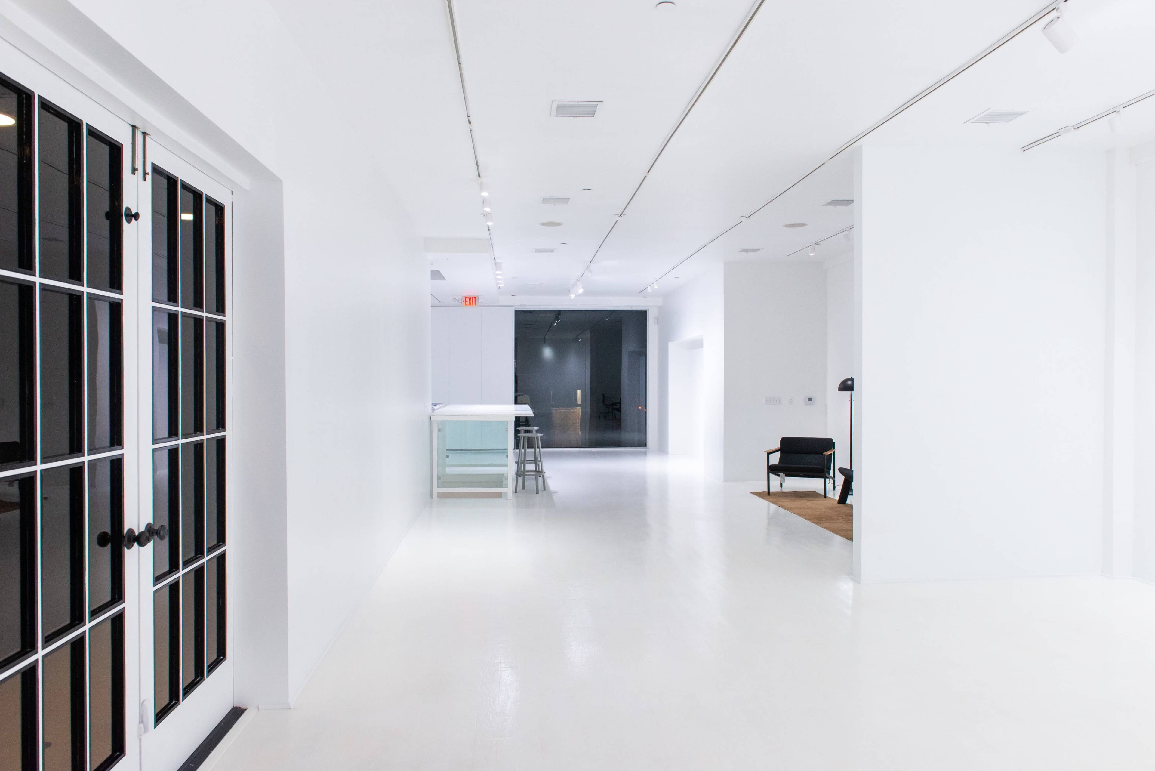 A spacious, primarily white interior with large windows, illuminated by overhead lights and featuring minimal furniture along the walls.