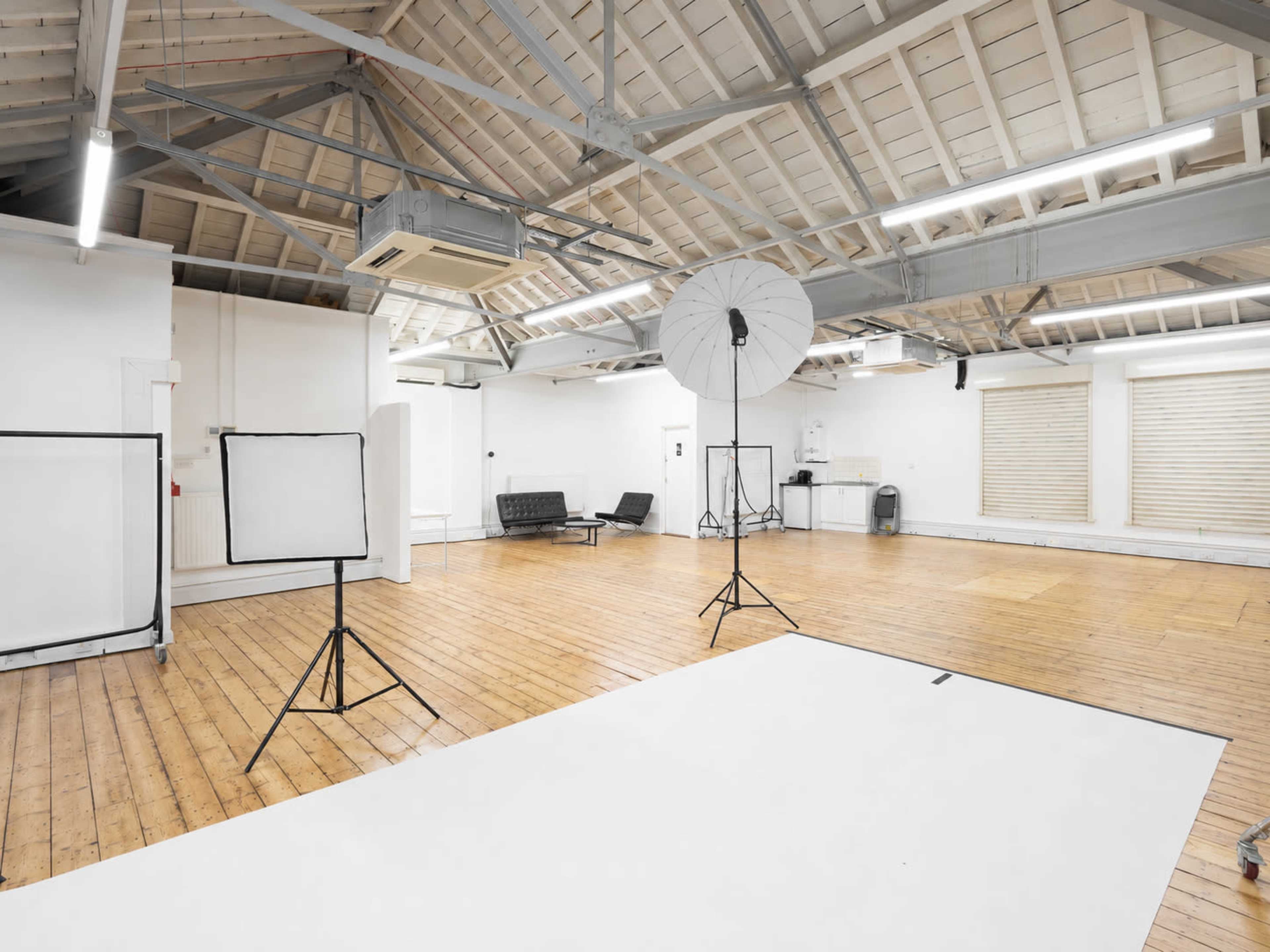 The image shows a spacious photography studio with wooden flooring, white walls, and various light equipment set up, including a large umbrella light and two backdrop stands.