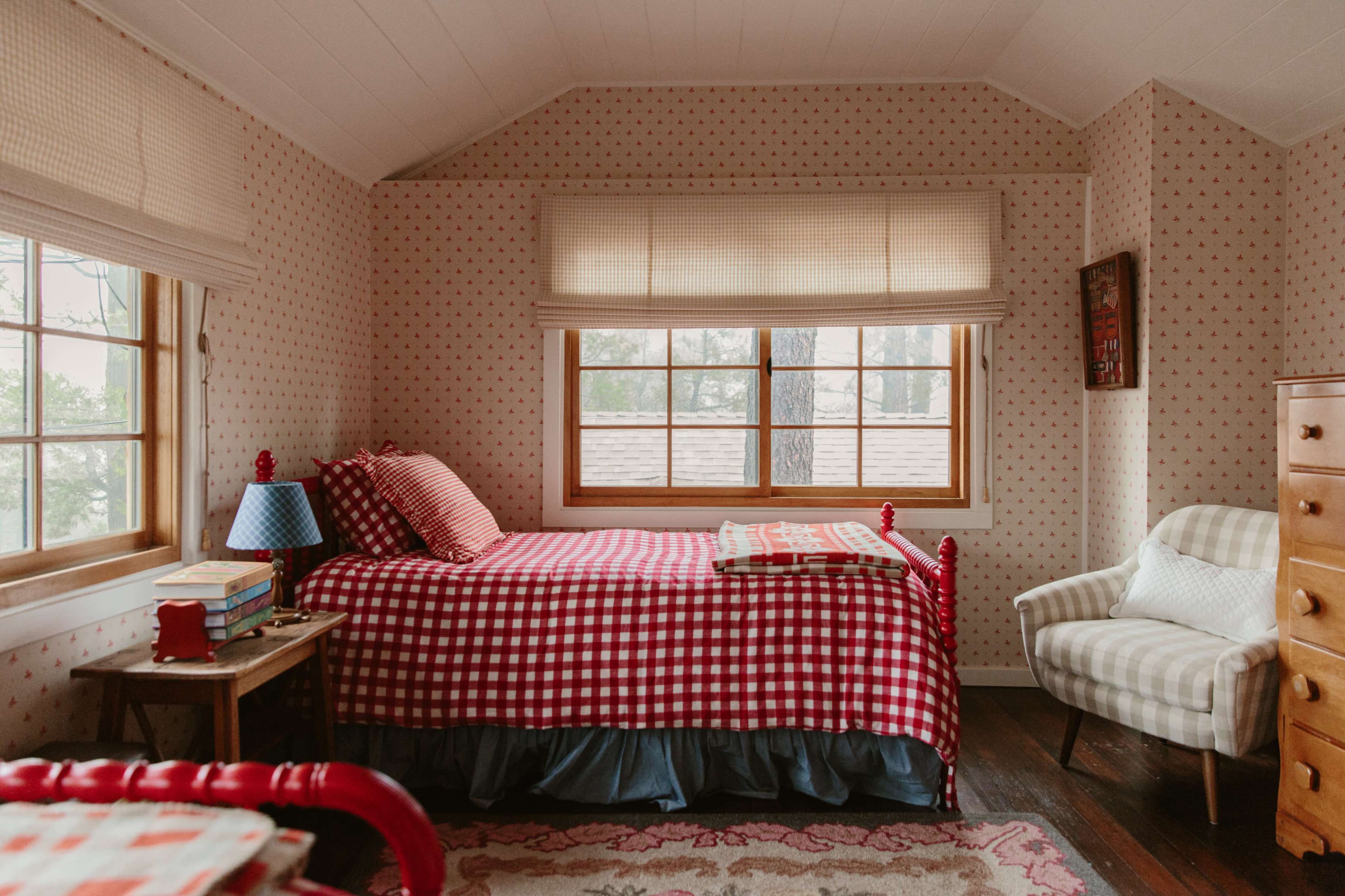 The image shows a cozy bedroom with a plaid bedspread, a small table with a blue lamp, and a comfortable chair near a window with natural light.