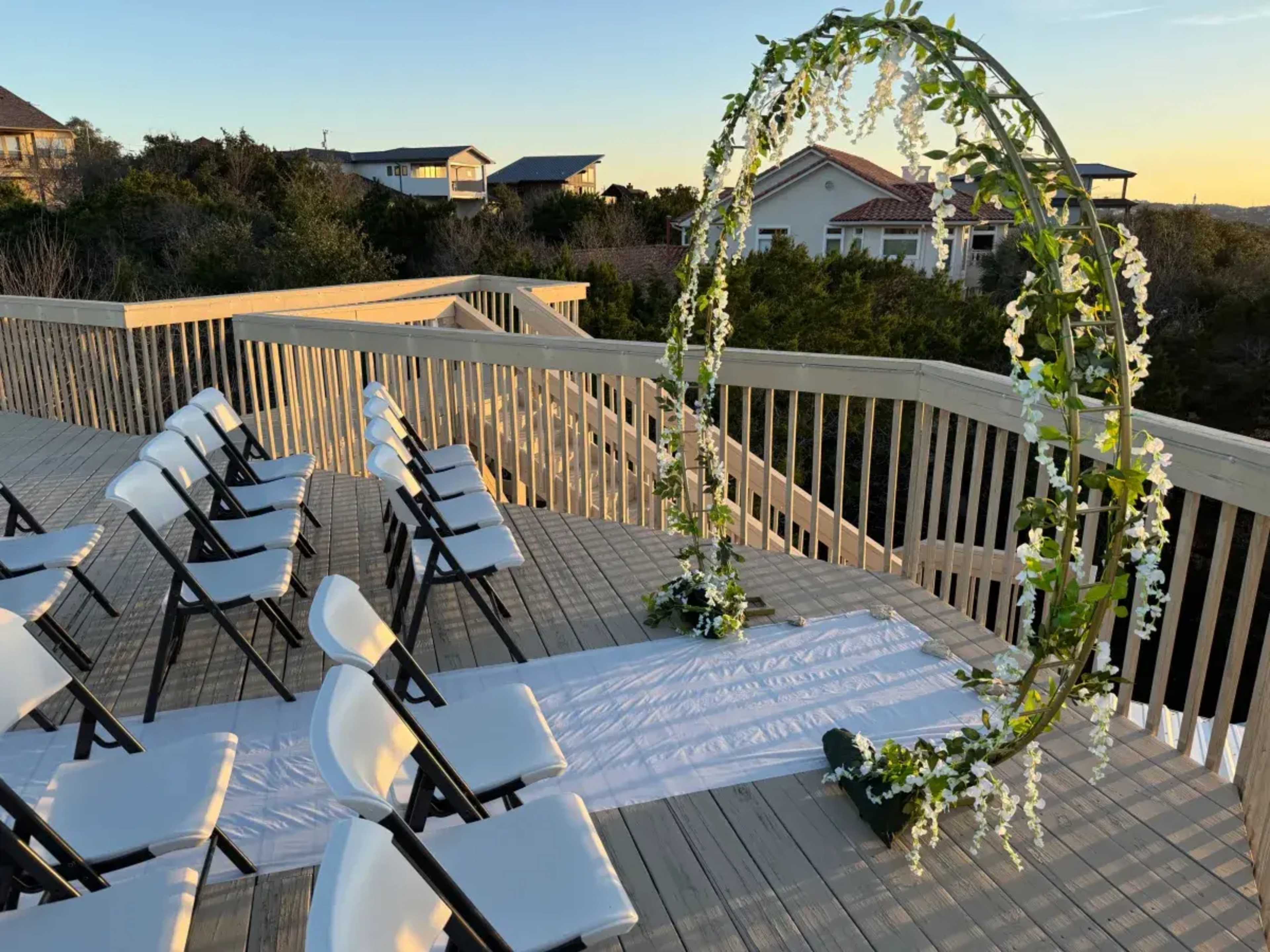 A circular floral arch is set up on a wooden deck, with rows of folding chairs facing it, overlooking a scenic view during sunset.