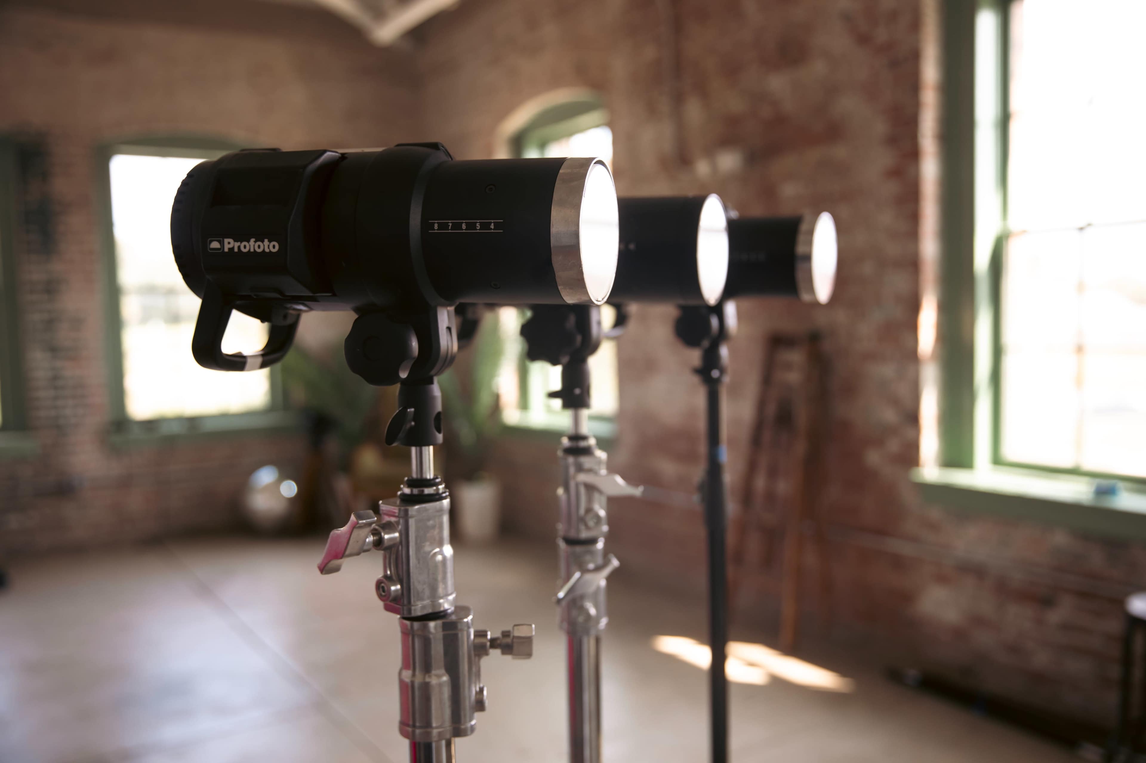 Three Profoto studio lights are mounted on stands in an industrial-style room with large windows and exposed brick walls.