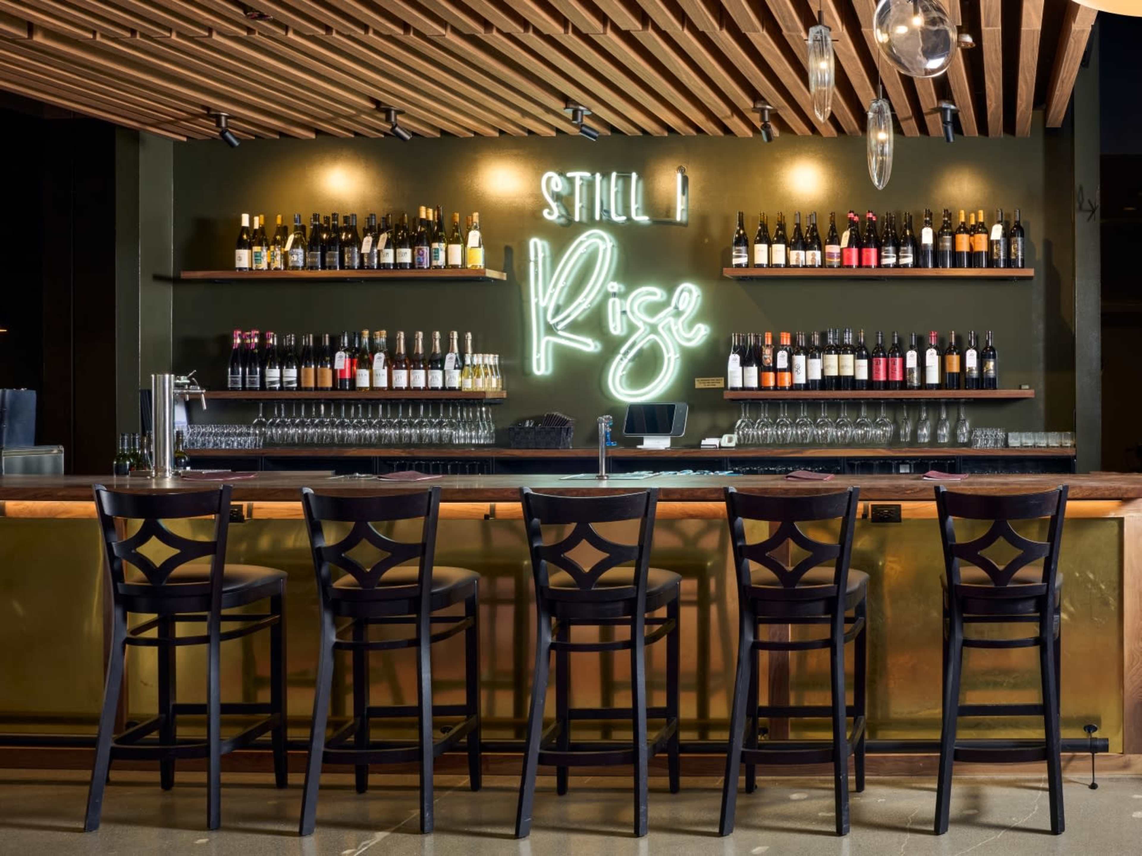 A sleek bar with a wooden counter features numerous bottles lined on shelves behind it, illuminated by a neon sign that reads "Still I Rise."