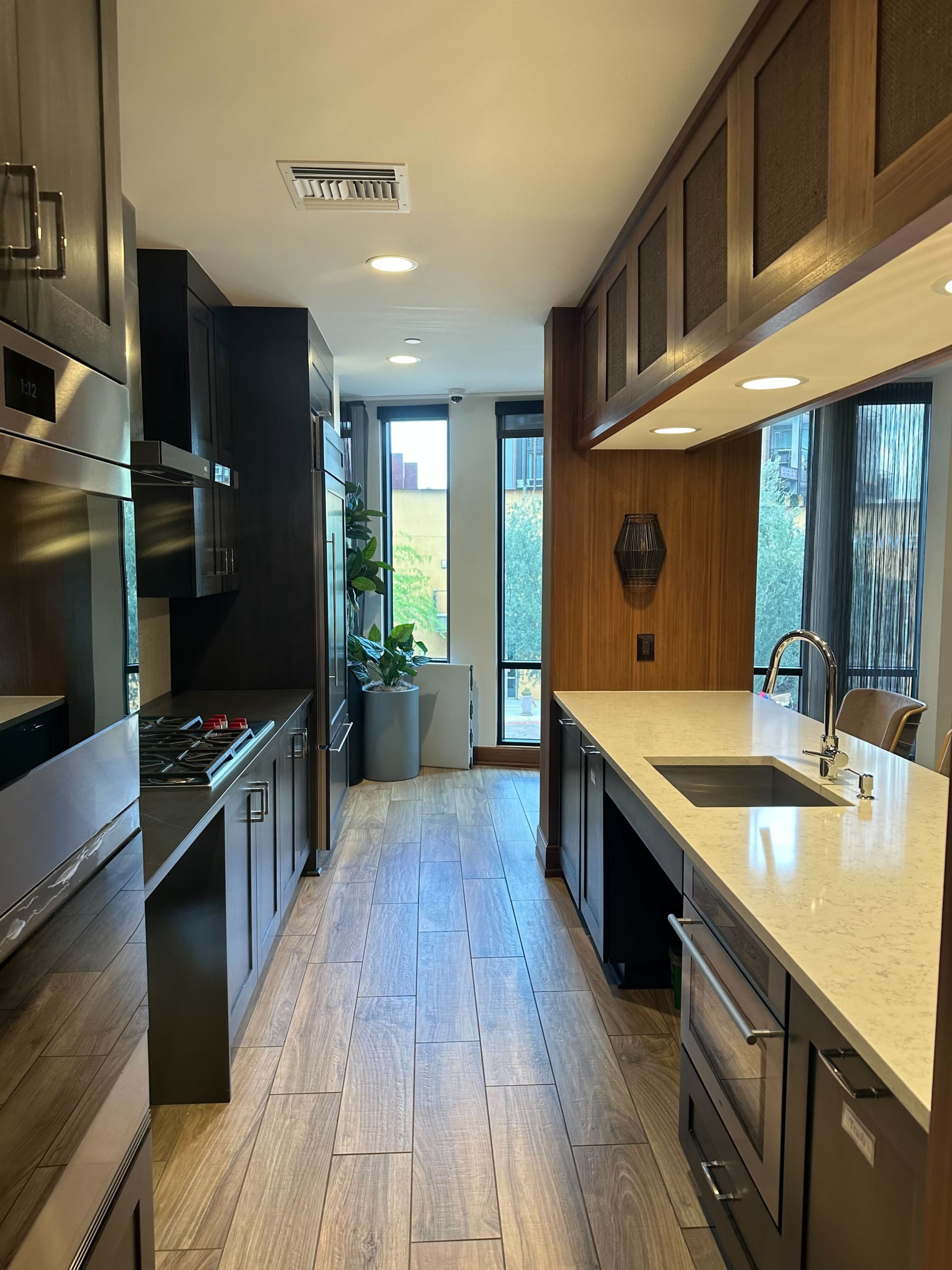 The kitchen features sleek black cabinetry, stainless steel appliances, and a white countertop, with a view of greenery through large windows.