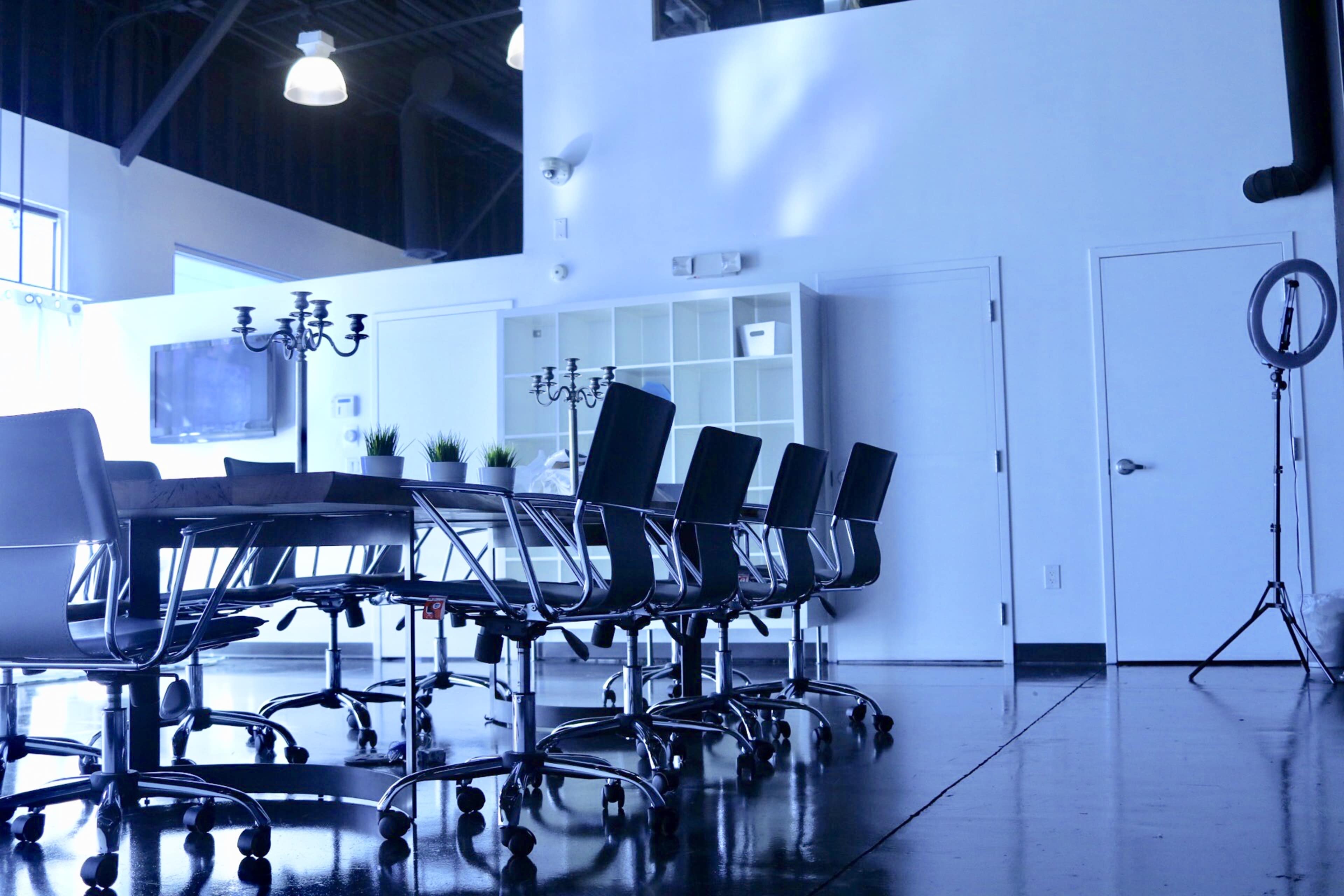 A long conference table with black chairs is placed in a bright, minimalist meeting room featuring a television and a shelf against the wall.