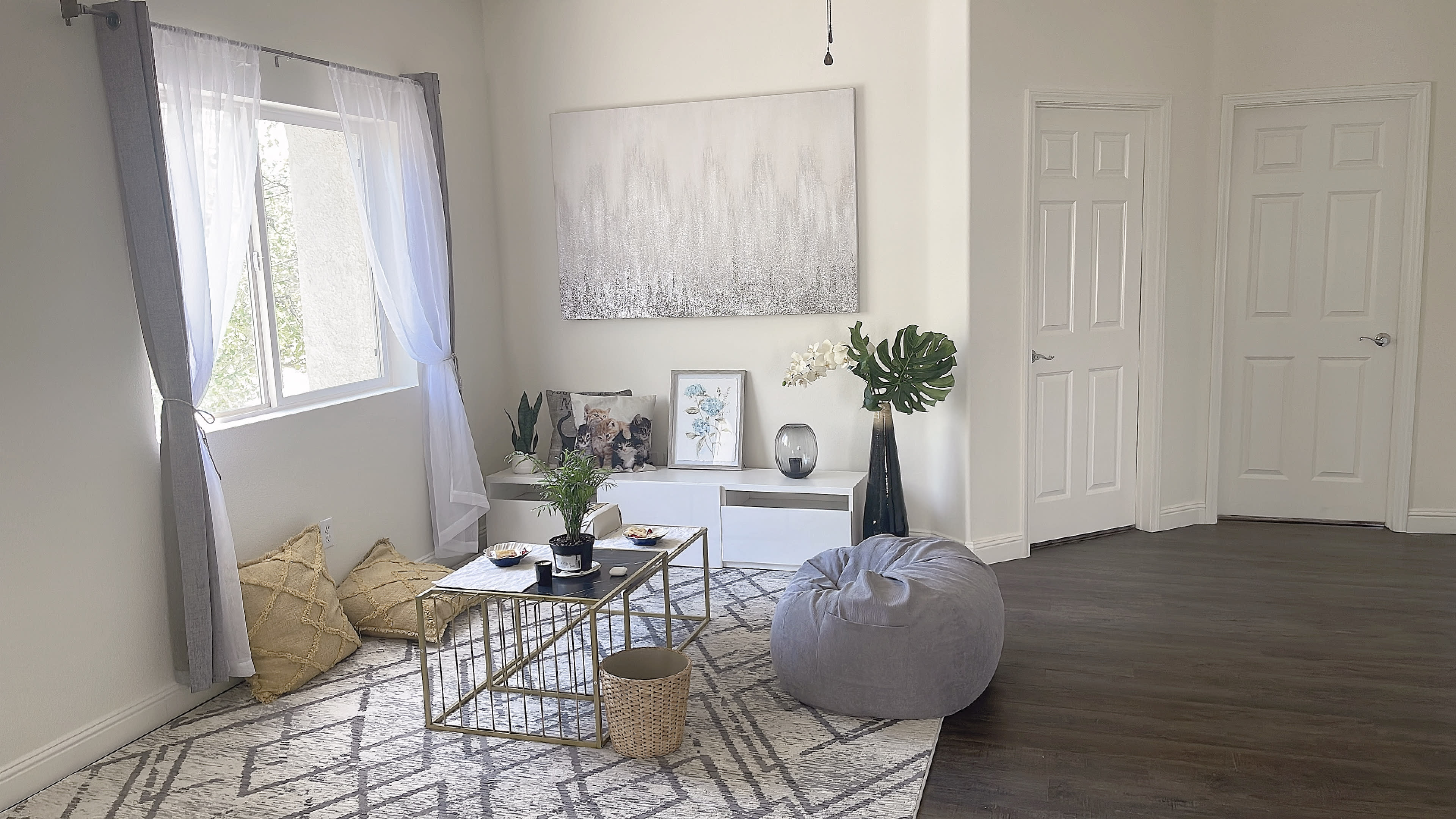A modern living room with a geometric rug, a low coffee table adorned with decor, a bean bag chair, and large windows allowing natural light to enter.