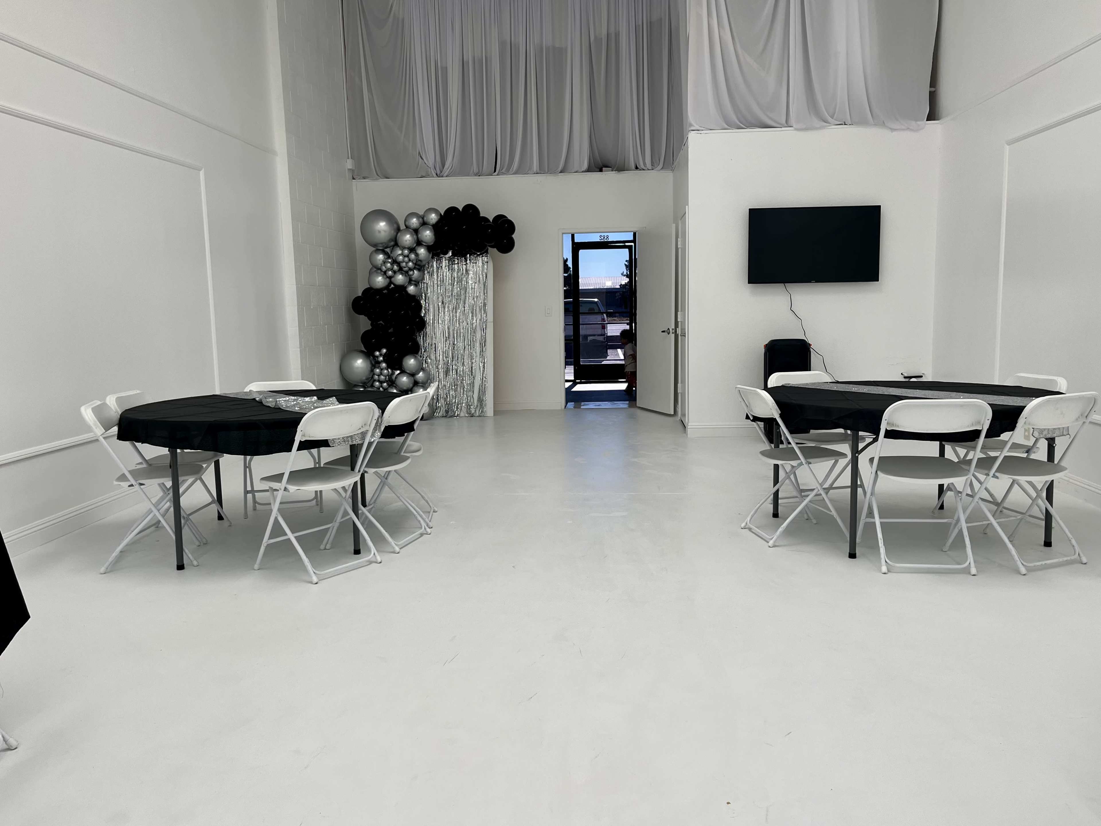 The image shows a spacious, white-walled room with black and white tables set for an event, featuring a decoration of black and silver balloons in one corner and a television mounted on the wall.