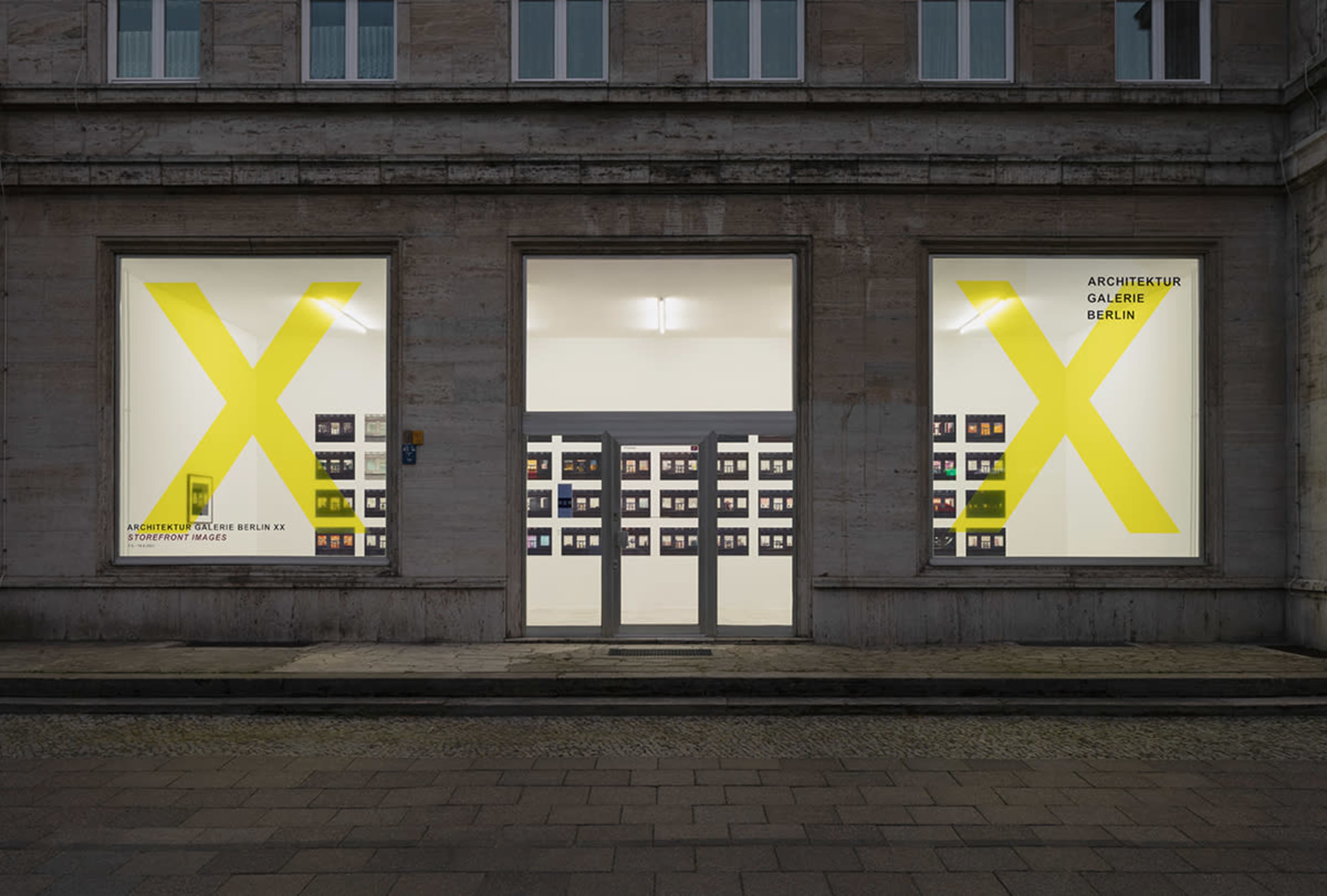 A gallery exterior in Berlin with large yellow letters "X" on the windows and a collection of framed photographs displayed on the wall inside.