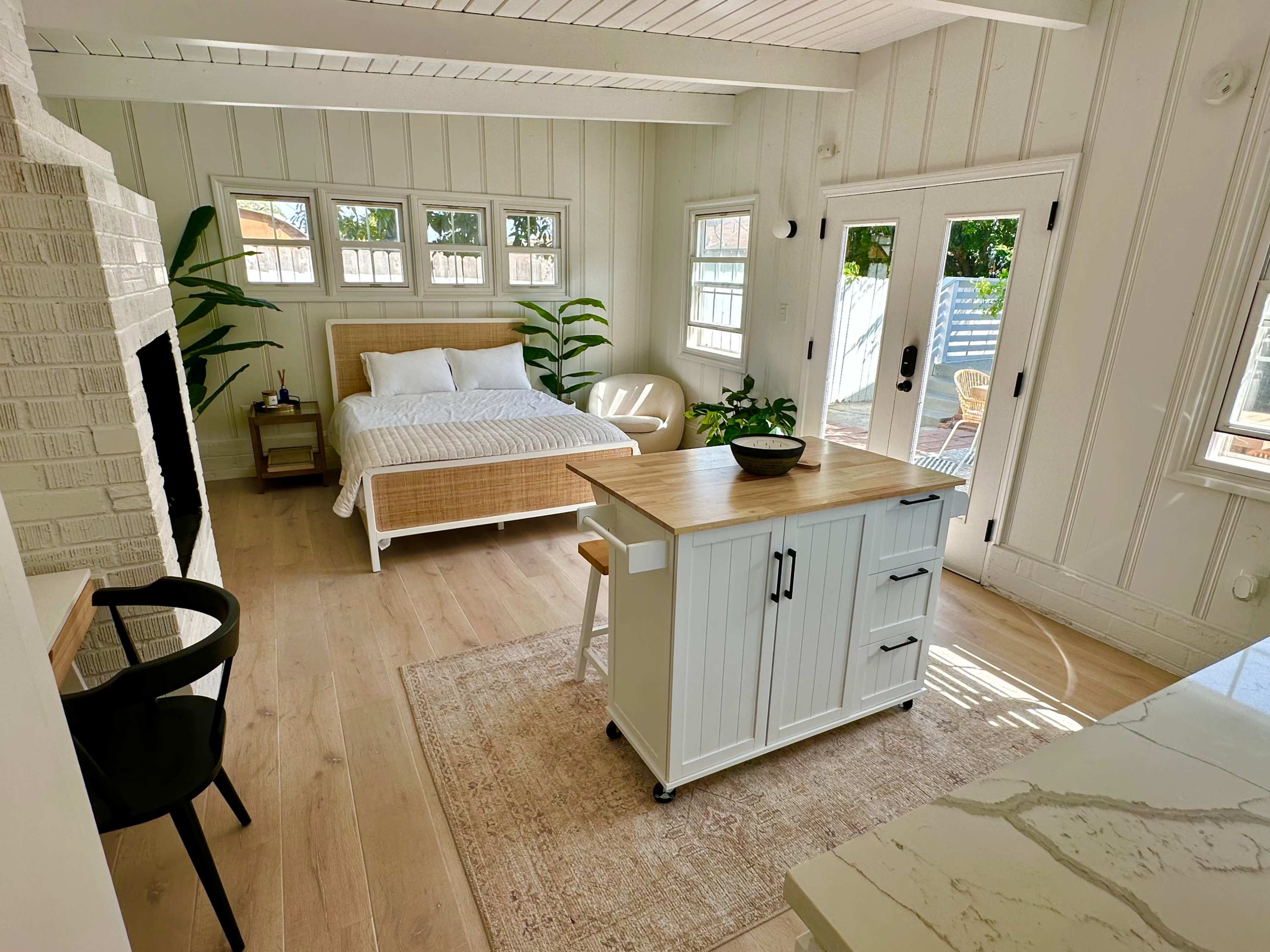 A bright, modern room features a bed, a small kitchen island, and large windows overlooking a patio with greenery.