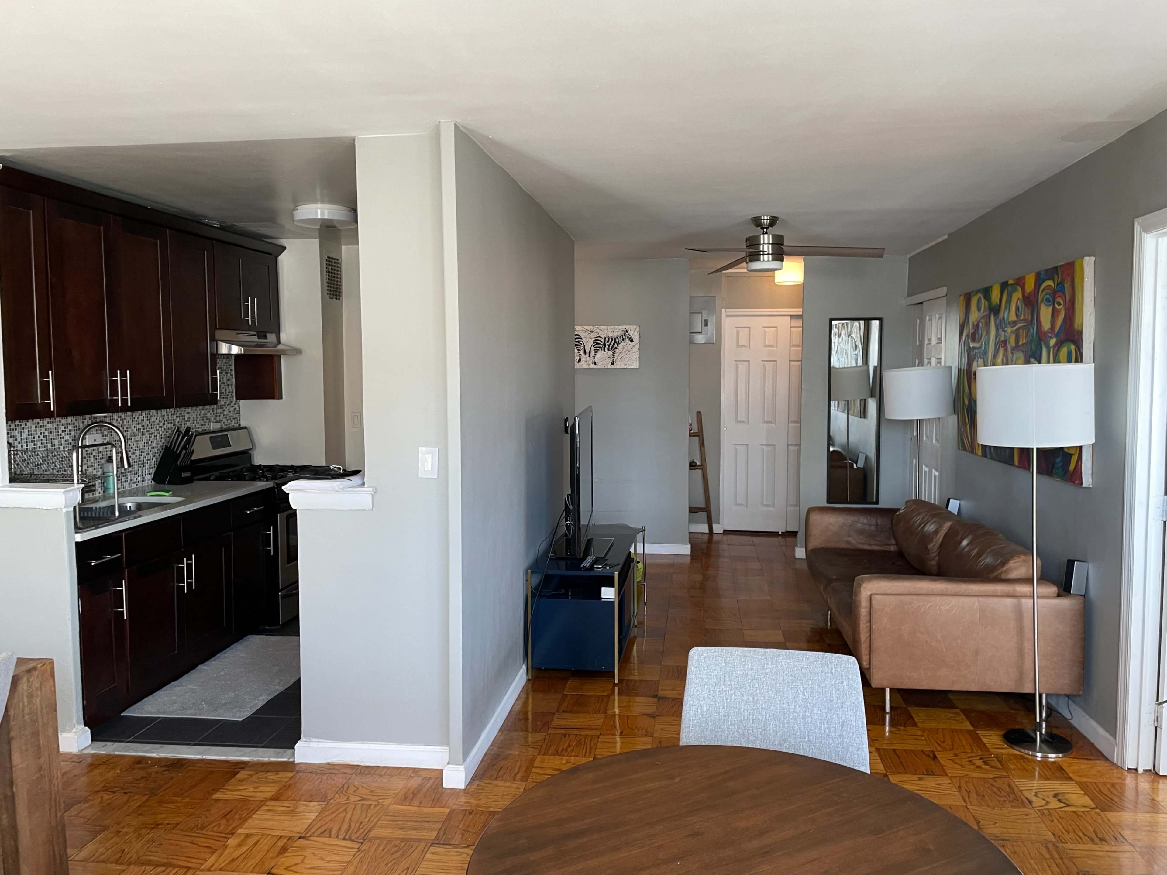 The image shows a modern open-concept living space that includes a kitchen area with dark wood cabinets, a dining table, a brown leather sofa, and a television.
