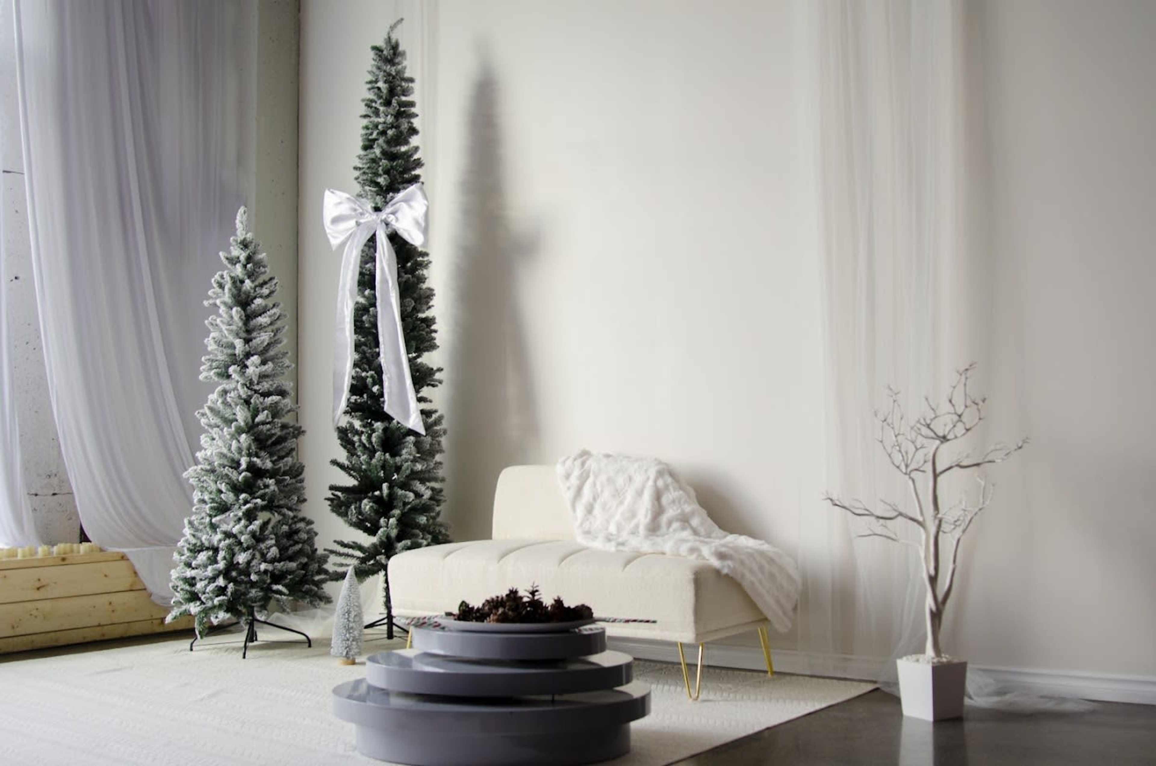 The image shows a minimalist interior with two artificial Christmas trees, a white sofa, and a decorative plant against a neutral wall.