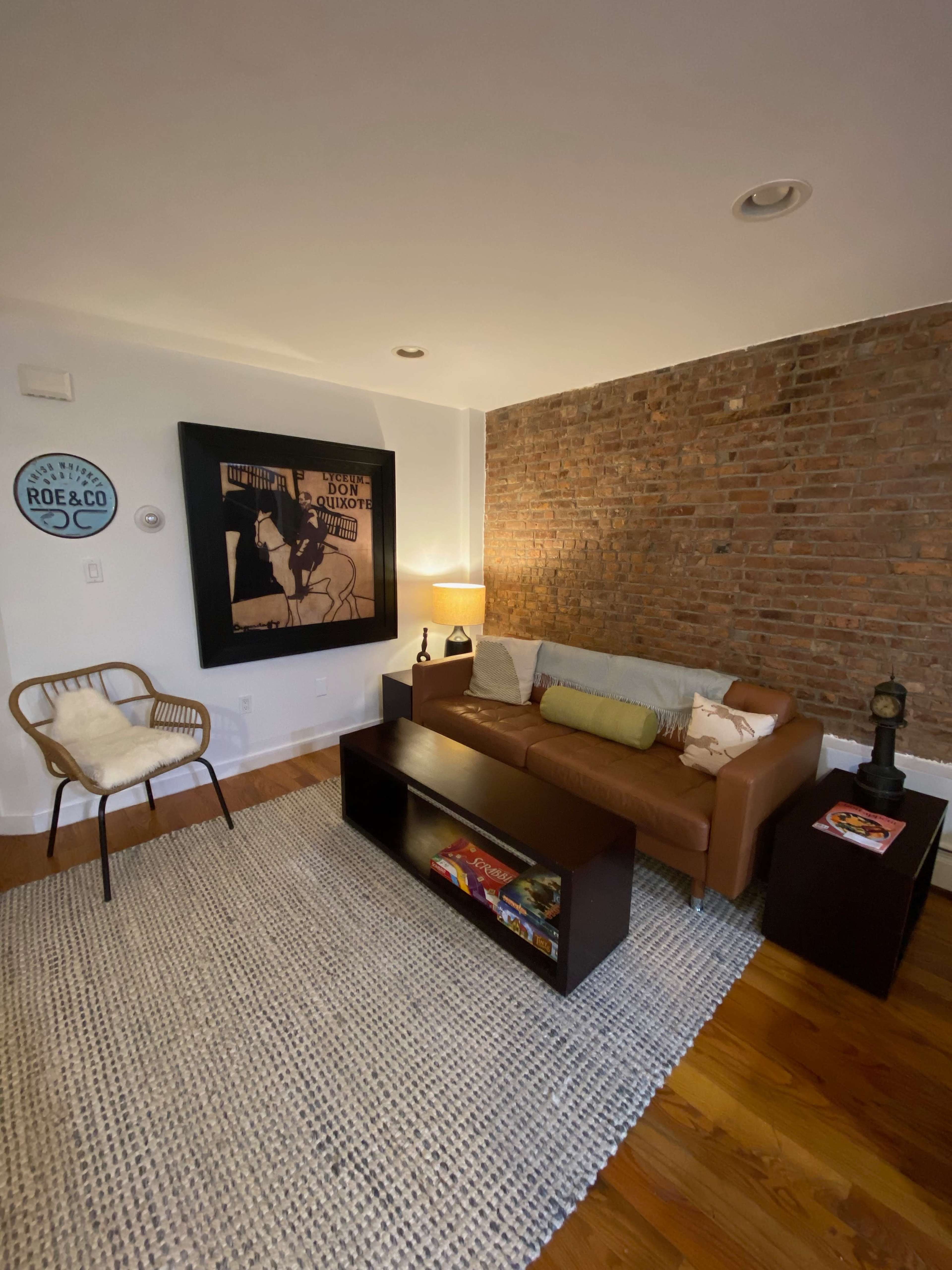 A cozy living room with a brown leather sofa, a wooden coffee table, a chair, a floor lamp, and a brick wall adorned with a vintage poster.