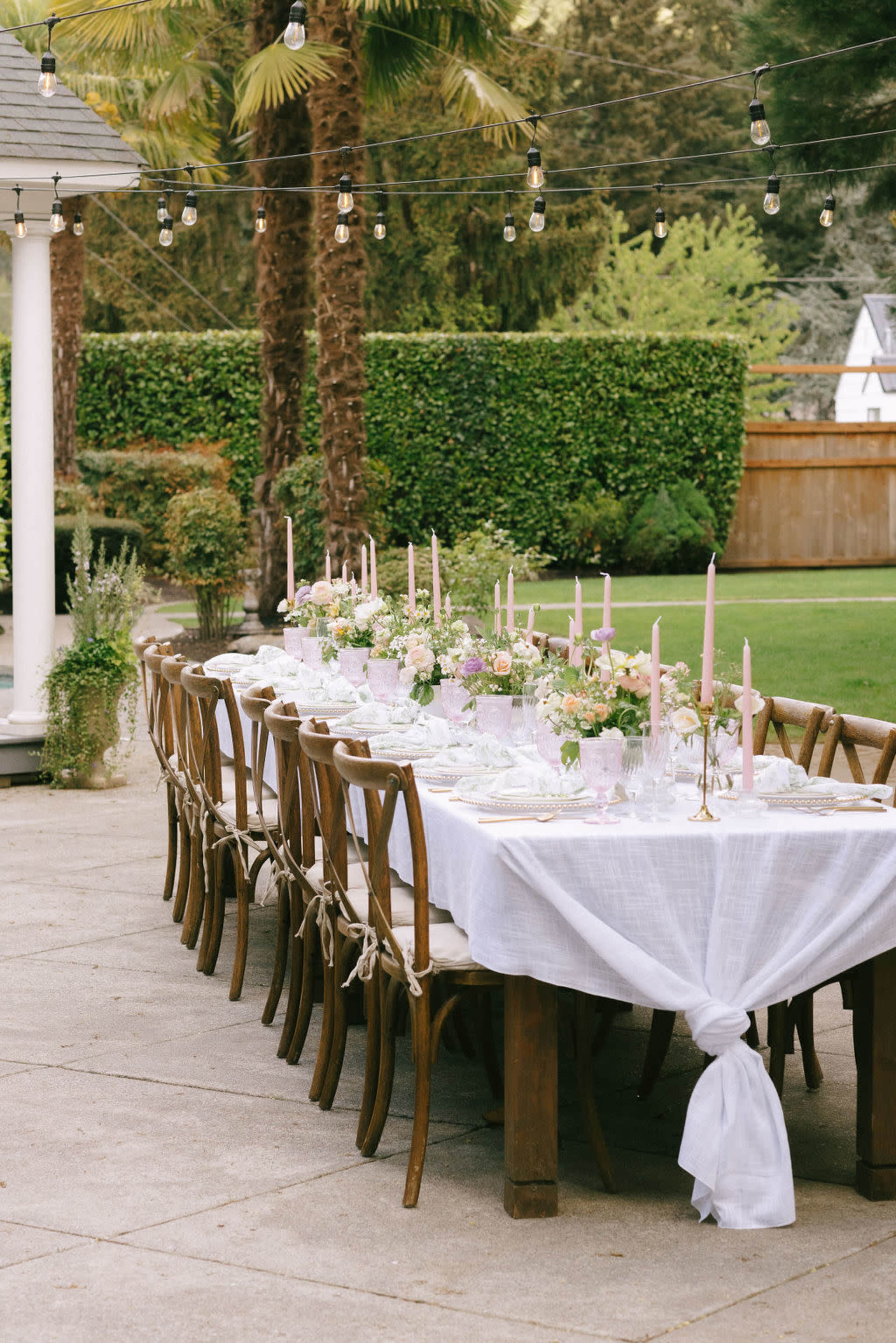 A long wooden dining table is set outdoors, adorned with flowers and candles, under string lights amidst greenery.