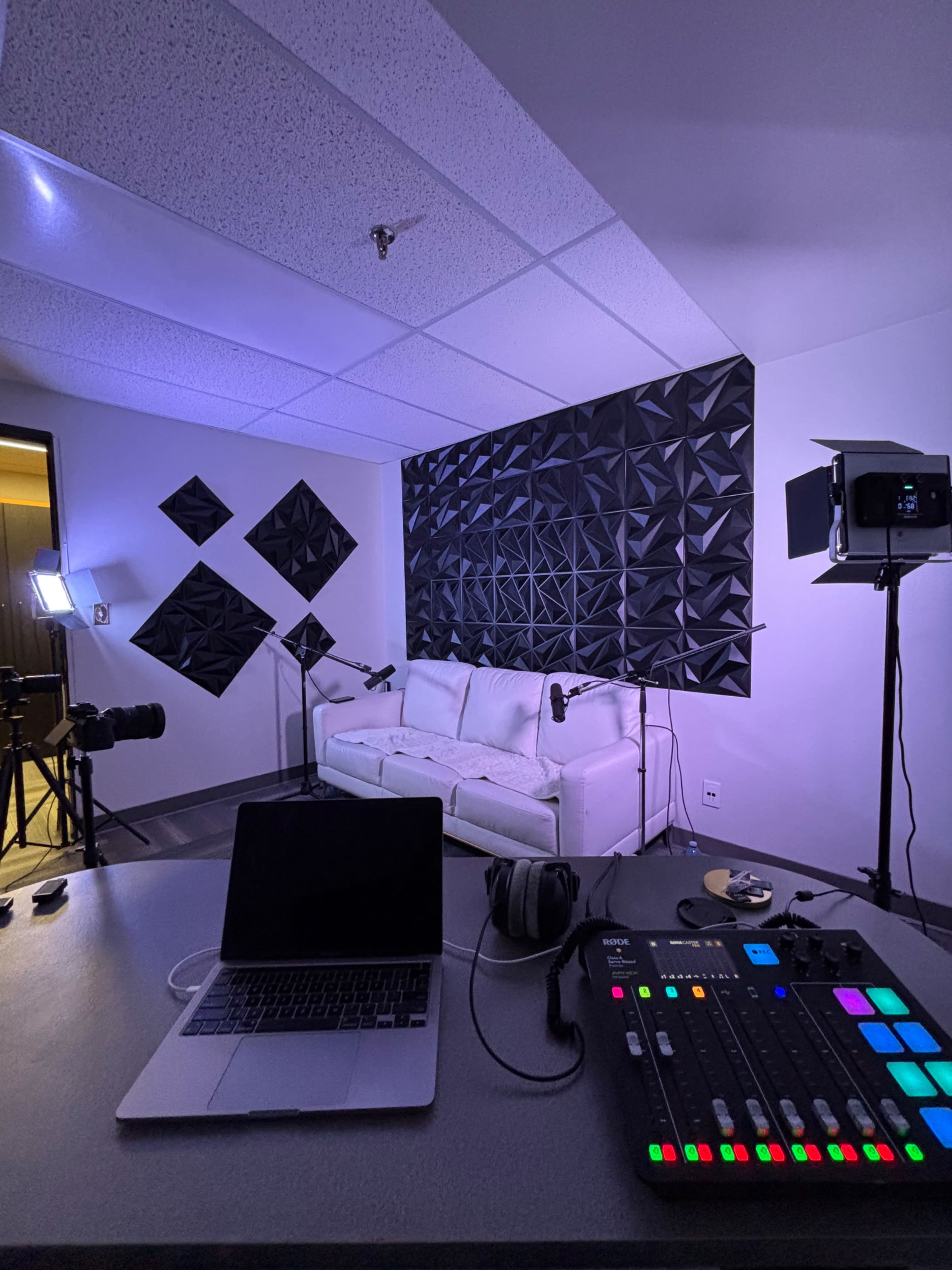 The image shows a modern studio setup with a white couch, soundproofing panels on the wall, a laptop, a sound mixing board, and camera equipment.