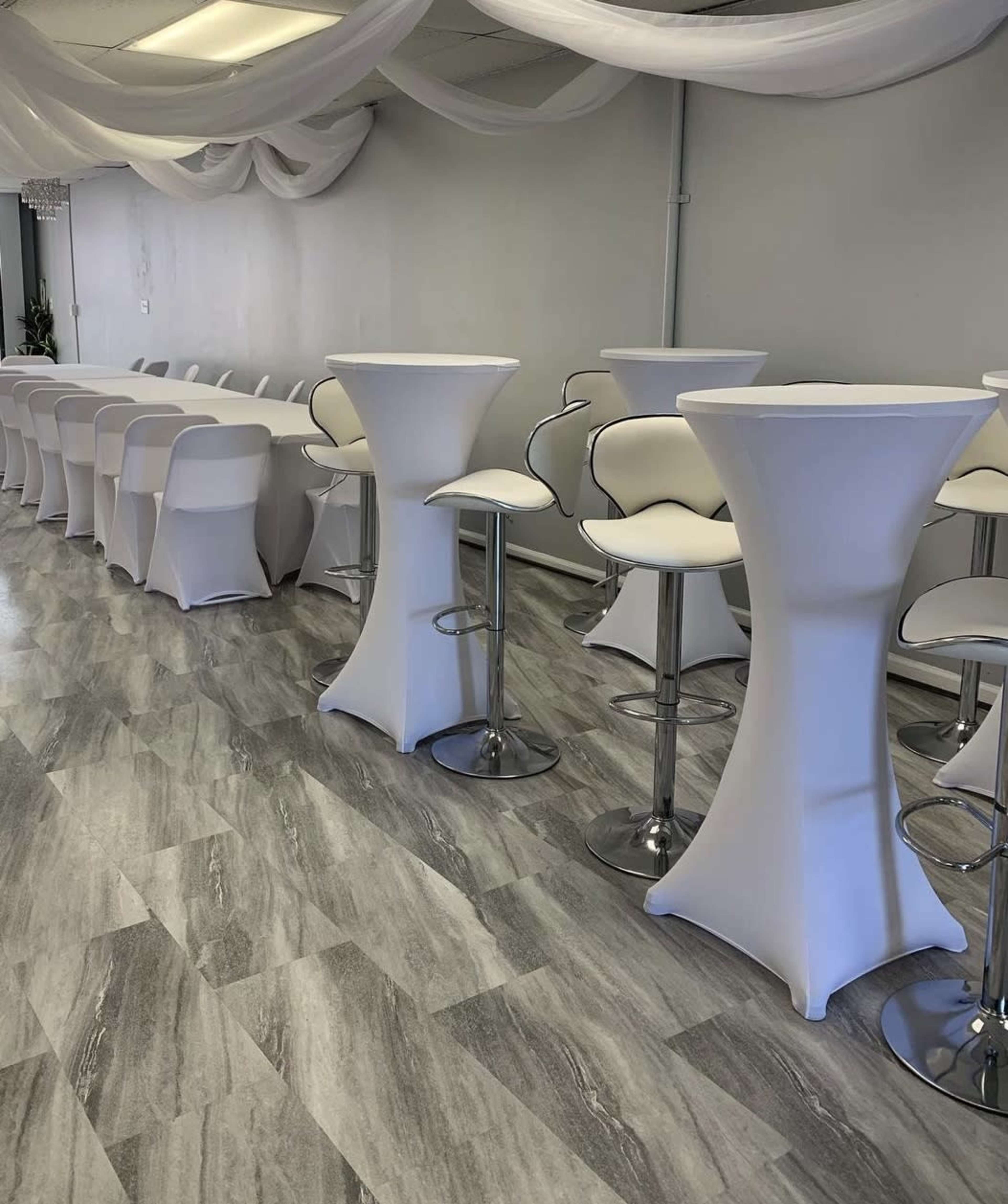 A spacious event room featuring white cocktail tables and high-backed stools arranged neatly along a light-colored wall.