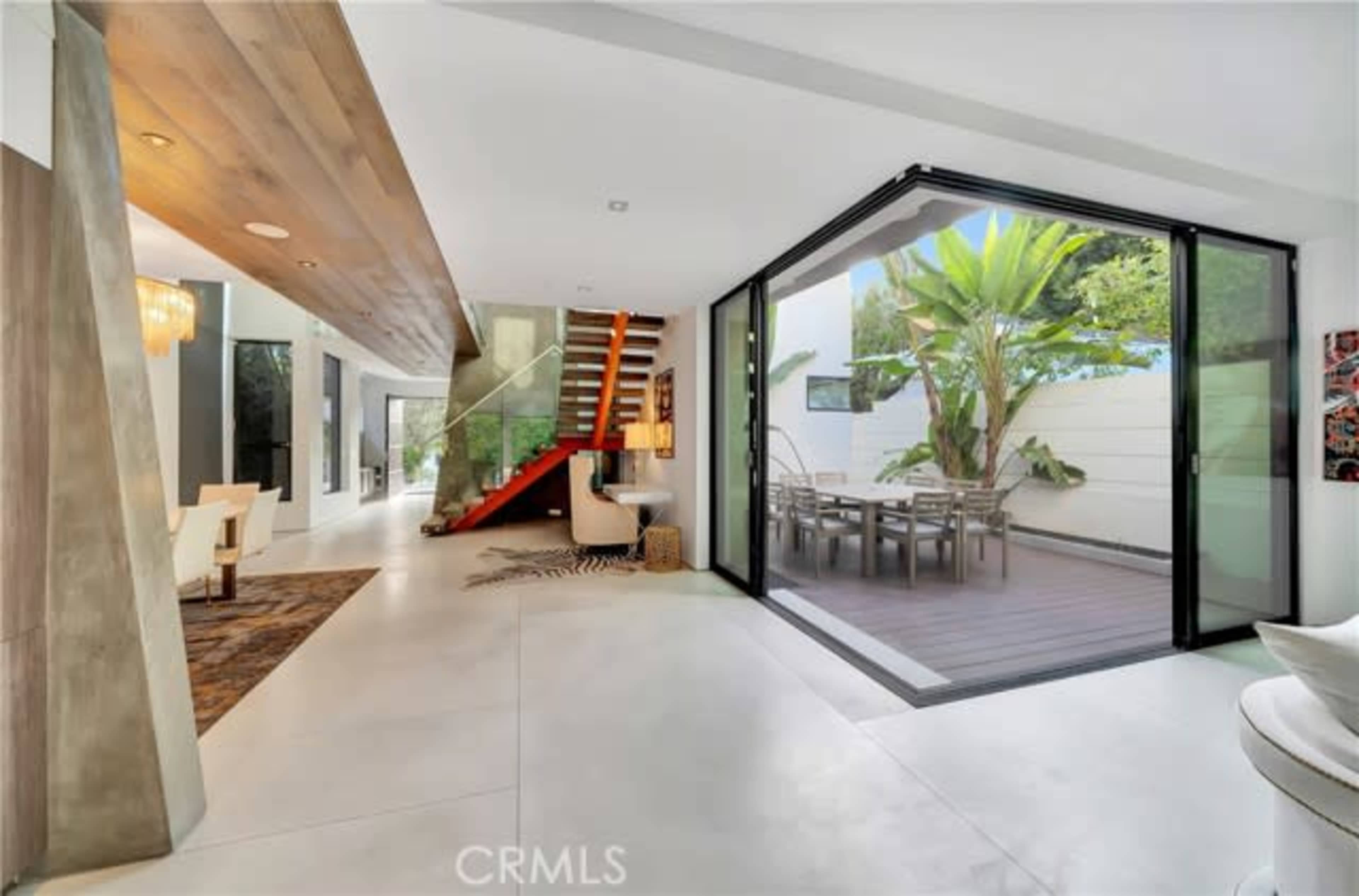 The image shows a modern interior space featuring a wooden staircase, large glass doors opening to an outdoor area, and a dining table surrounded by greenery.