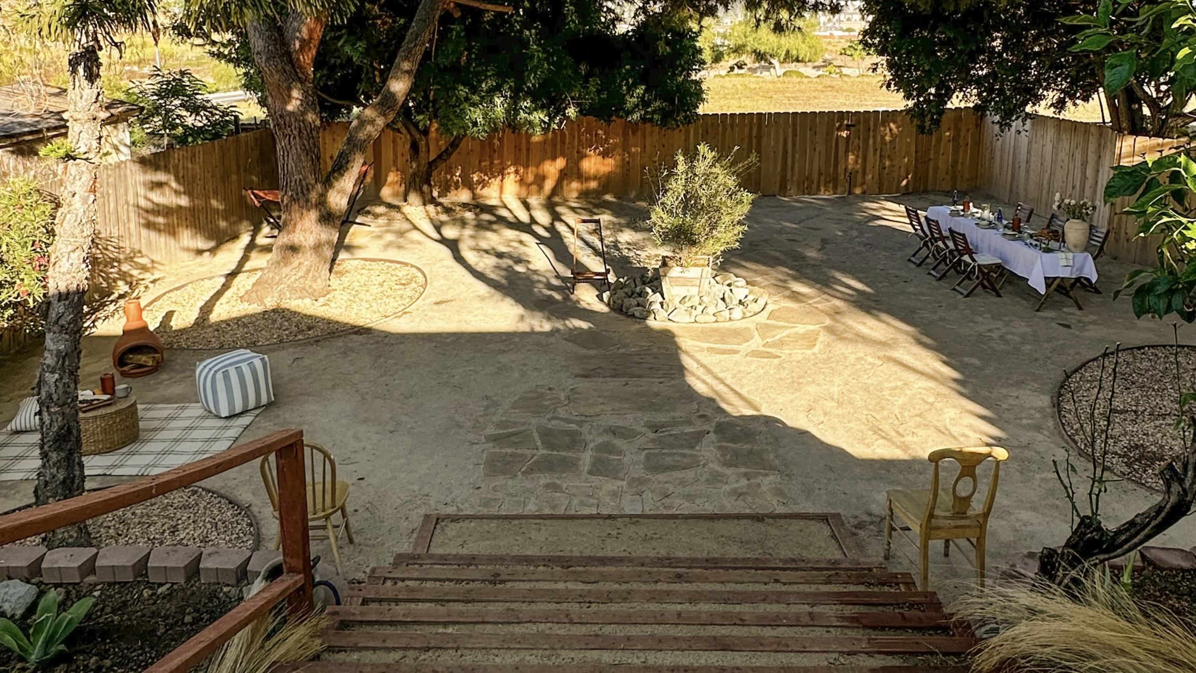 A spacious outdoor area with a stone pathway, a circular fire pit, several tables set for dining, and a wooden fence surrounding the scene.