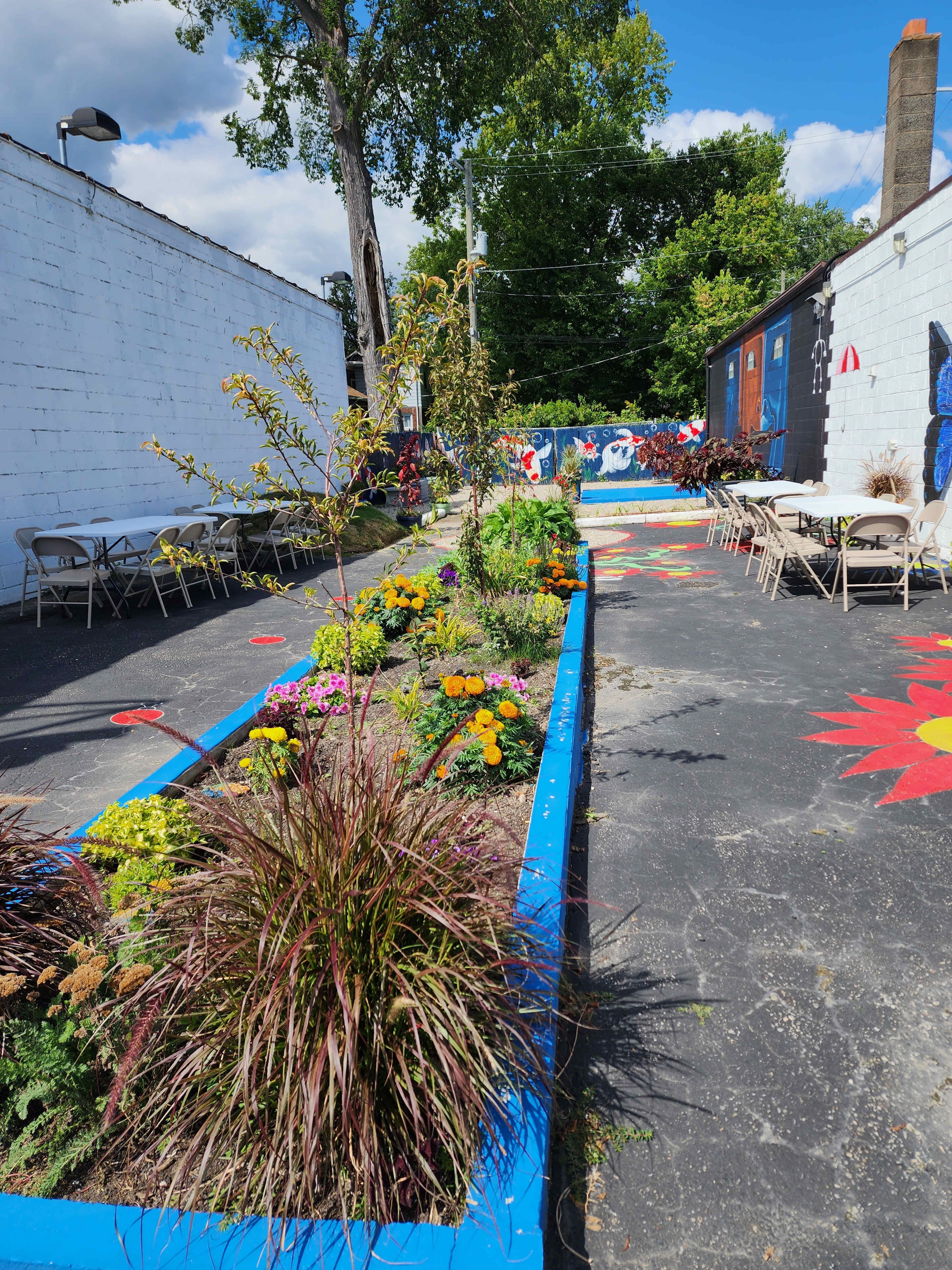 The image shows a colorful outdoor area with flower beds, seating arranged along the sides, and murals on the walls.