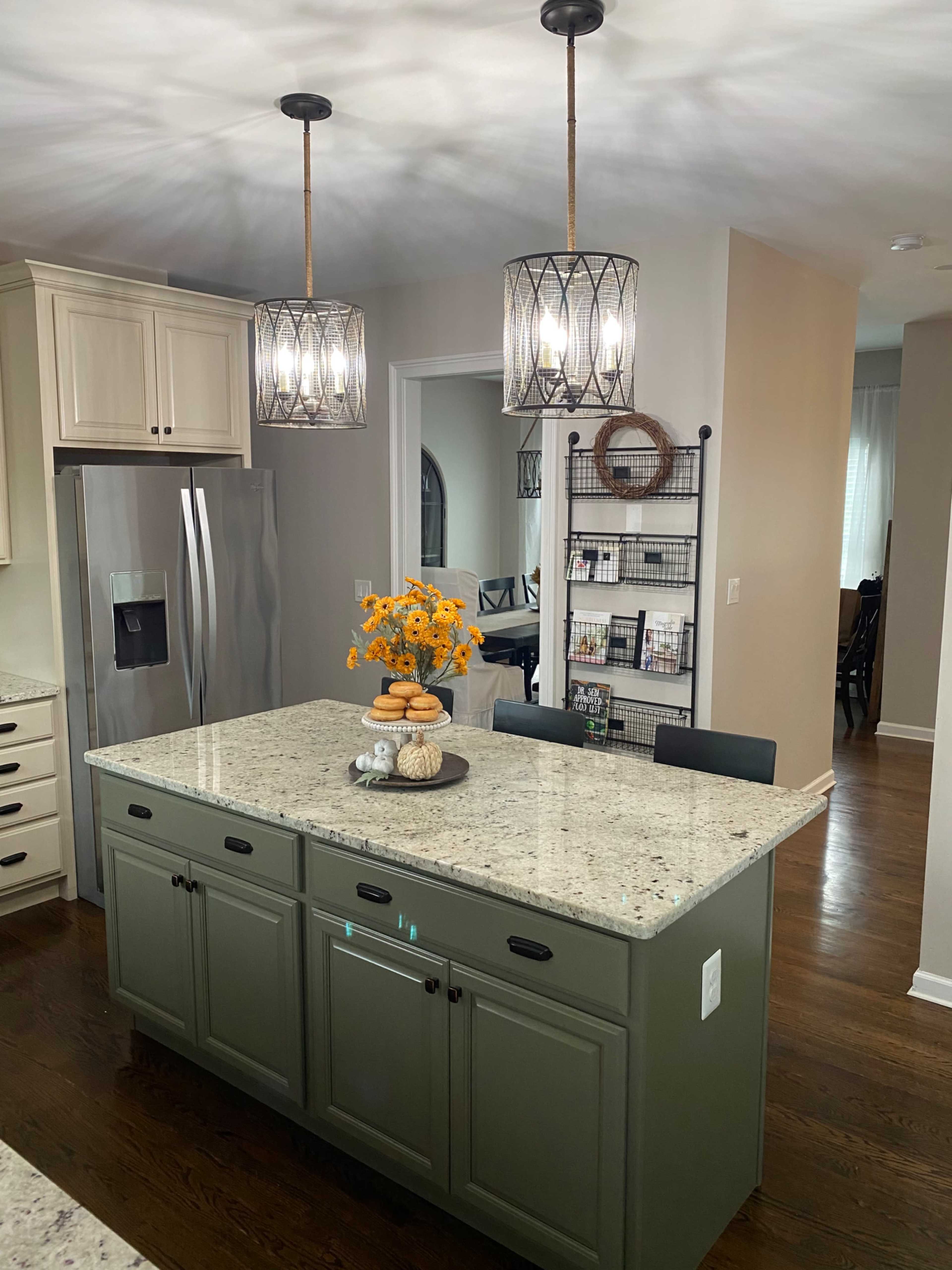 A modern kitchen features a large island with a green base and gray speckled countertop, surrounded by pendant lighting and a dining area in the background.
