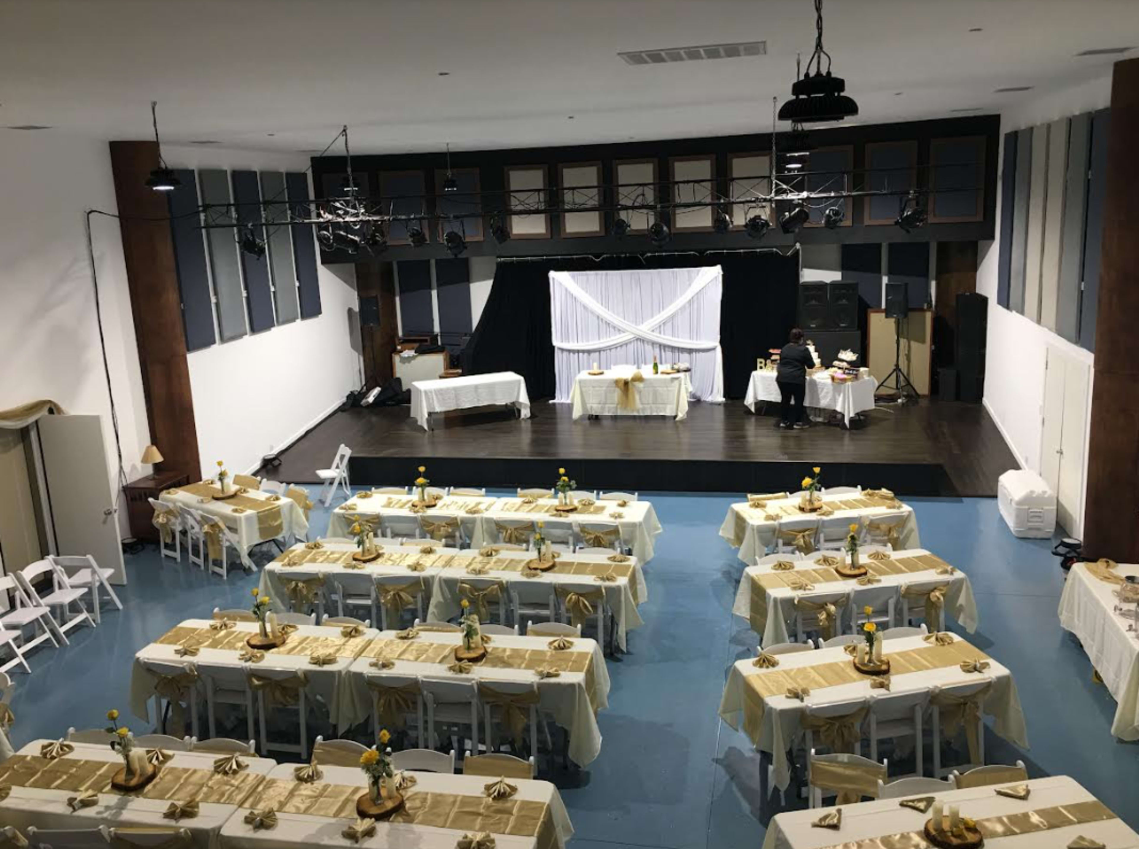 A spacious hall is set up for an event with neatly arranged tables decorated with yellow tablecloths and centerpieces.