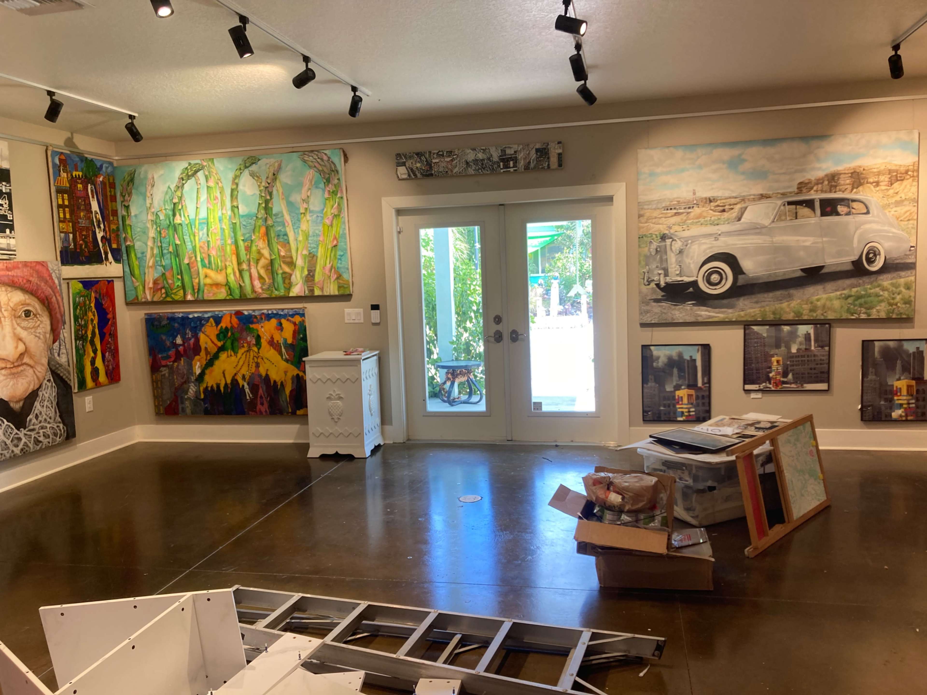 The image shows an art gallery with colorful paintings on the walls, a white cabinet, and scattered furniture in the center of the room.
