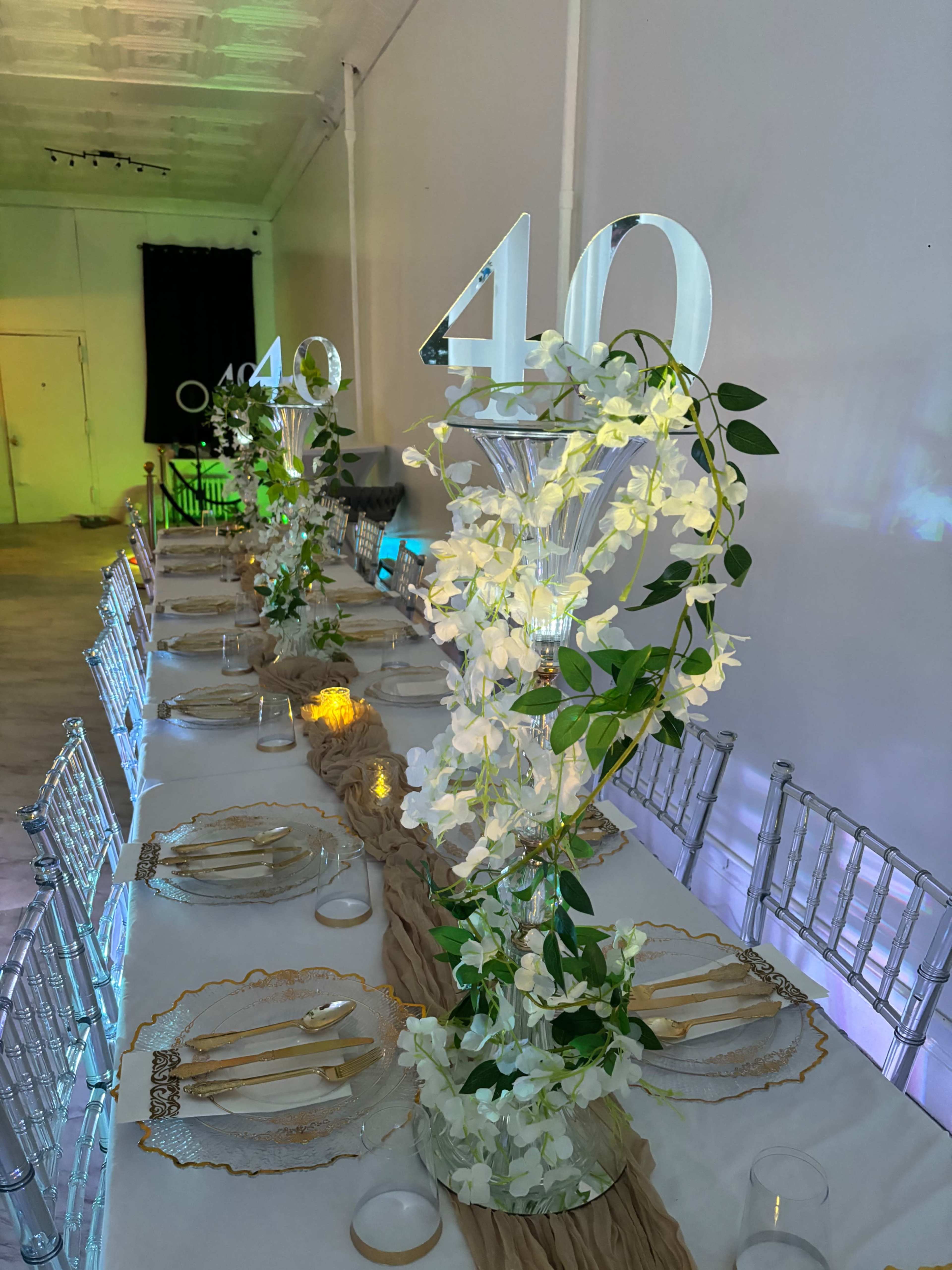 A long banquet table is decorated with floral centerpieces, elegant tableware, and illuminated number "40" displays.