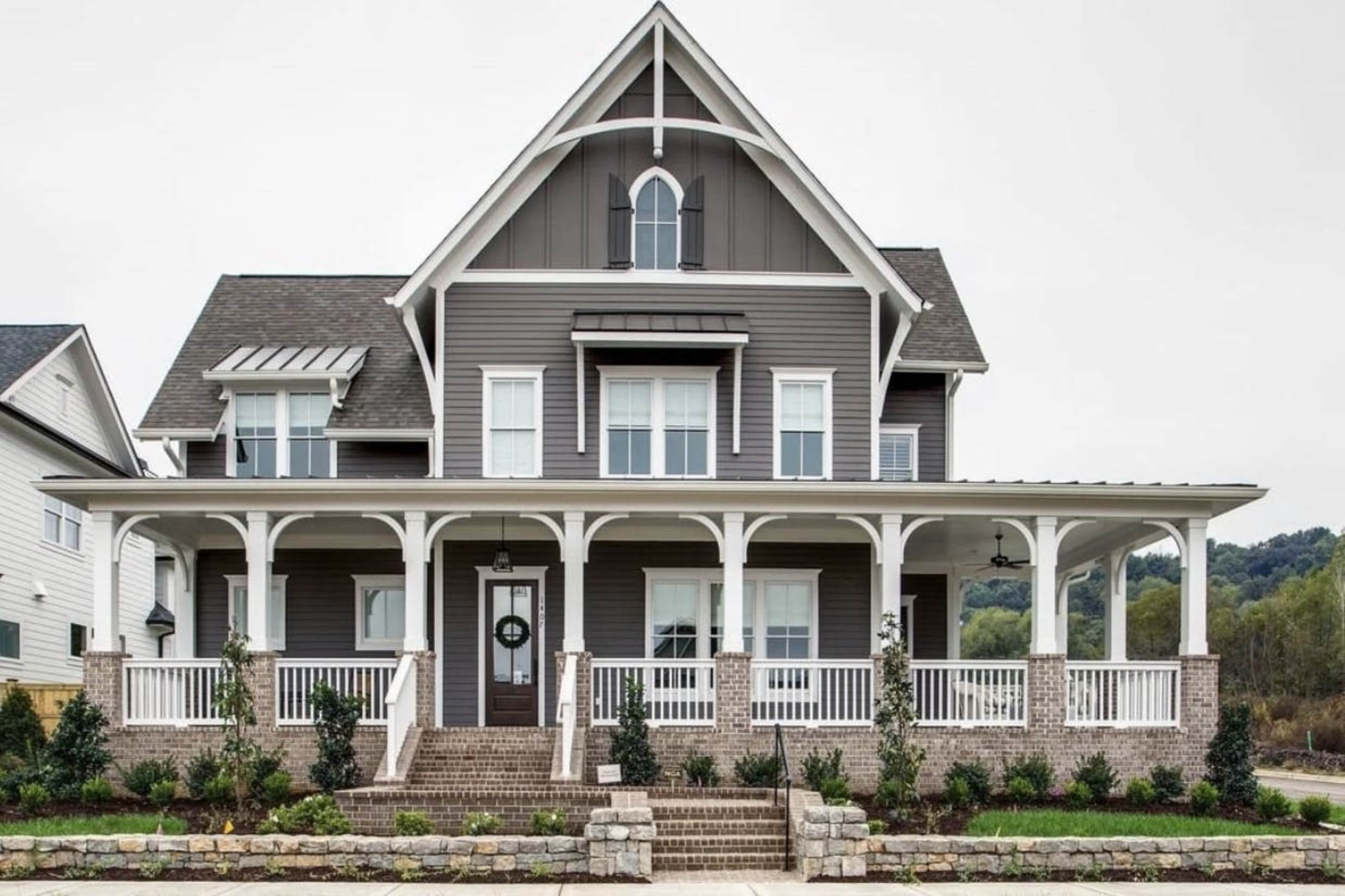 The image shows a large, gray two-and-a-half-story house with a front porch and white accents, surrounded by neatly trimmed landscaping.