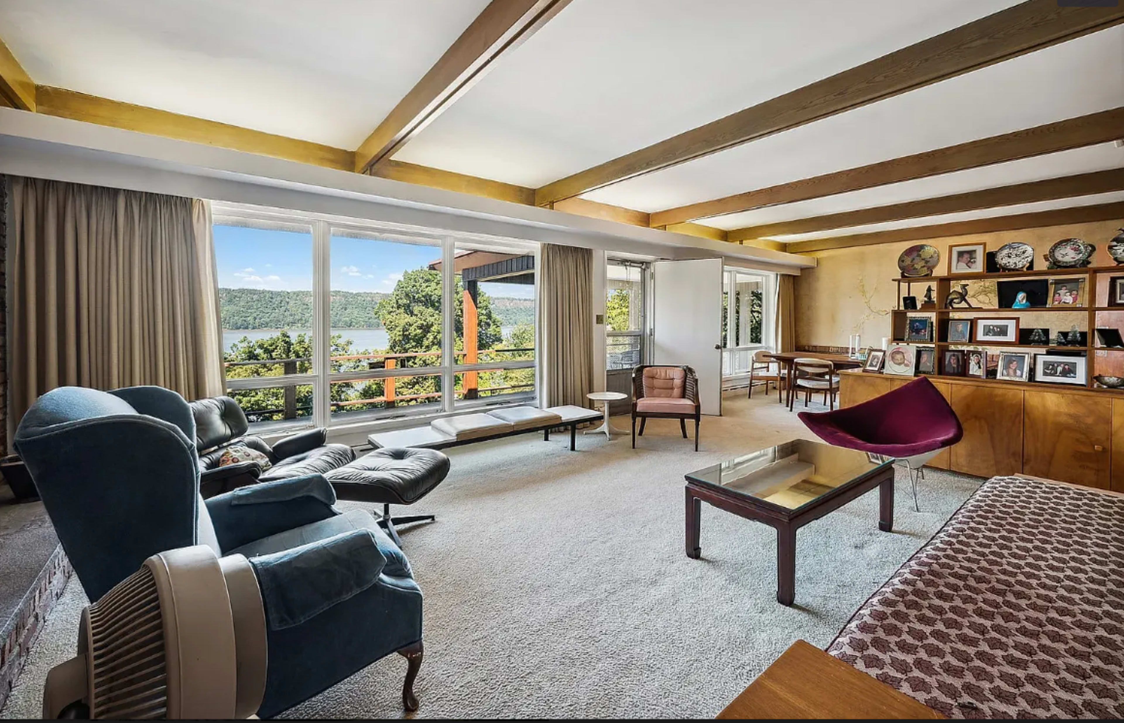 A modern living room with large windows overlooking a lake, featuring a mix of furniture including a blue armchair, a sleek black lounge chair, and a wooden shelving unit displaying decorative items.